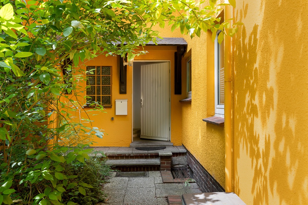 Eingangsbereich eines gelben Wohnhauses mit Pflanzen und offenstehender Haustür.
