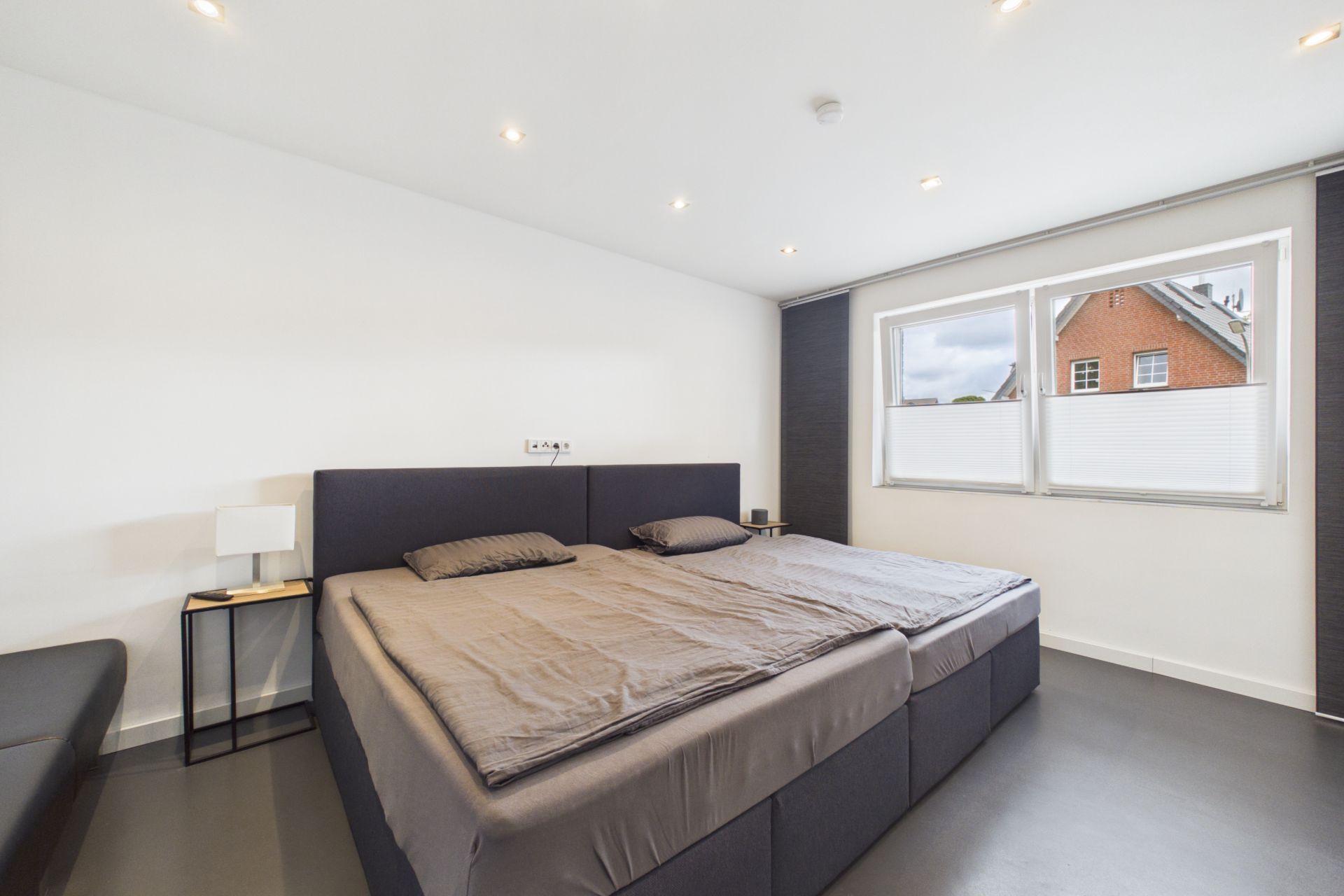 Modernes Schlafzimmer mit großem Doppelbett, Nachttischen und zwei Fenstern mit Sicht auf ein Haus.
