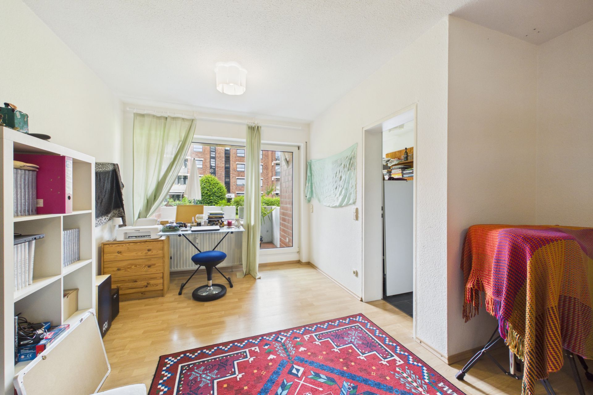 Helles Zimmer mit Schreibtisch, Regal, buntem Teppich und Zugang zum Balkon.