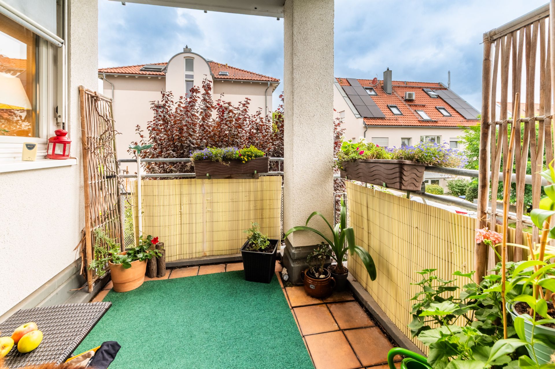 Balkon mit grünem Teppich, Pflanzen in Töpfen und Blumenkästen, Häuser im Hintergrund.