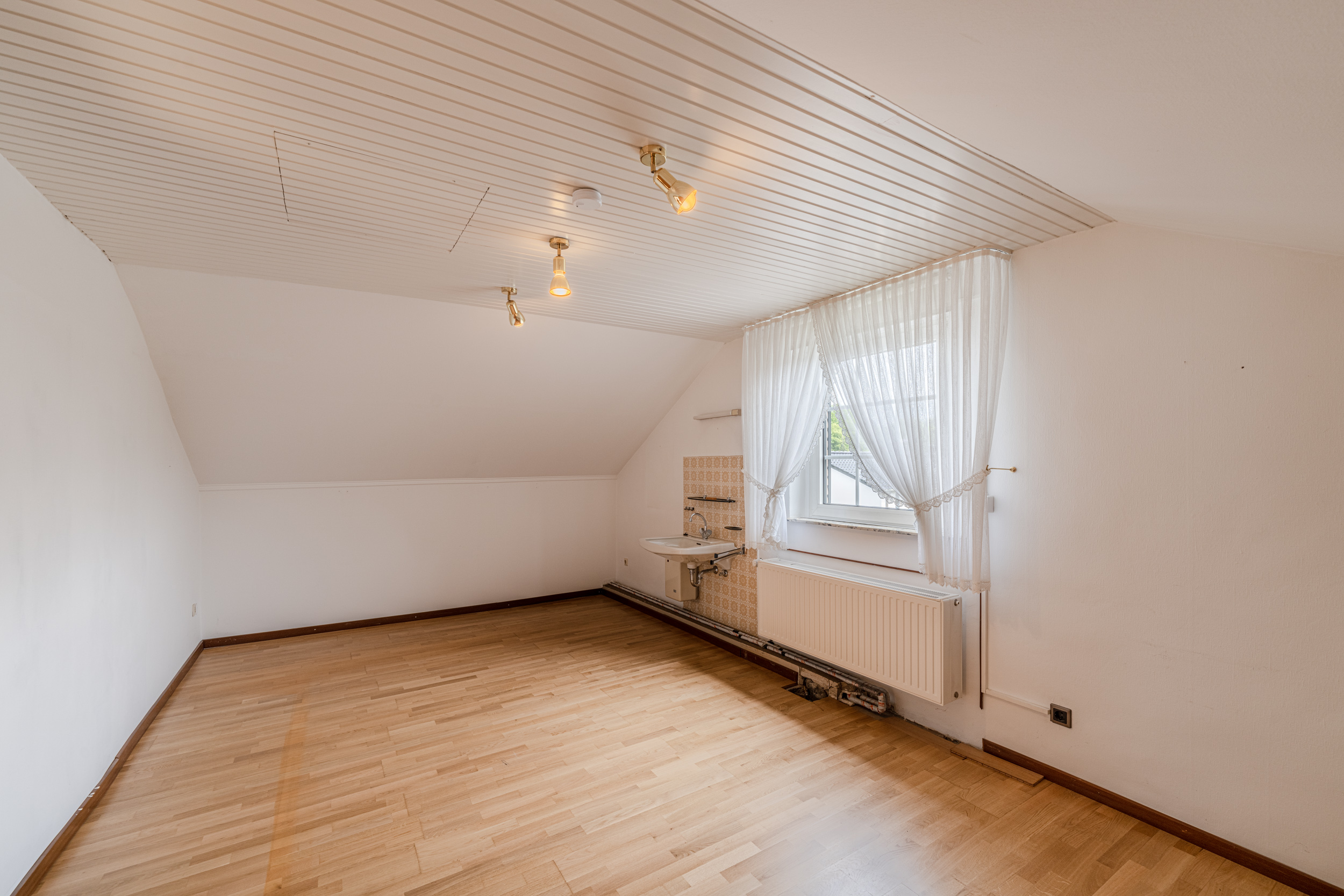 Leeres Zimmer mit Holzboden, Fenster mit Gardinen und Waschbecken an der Wand.