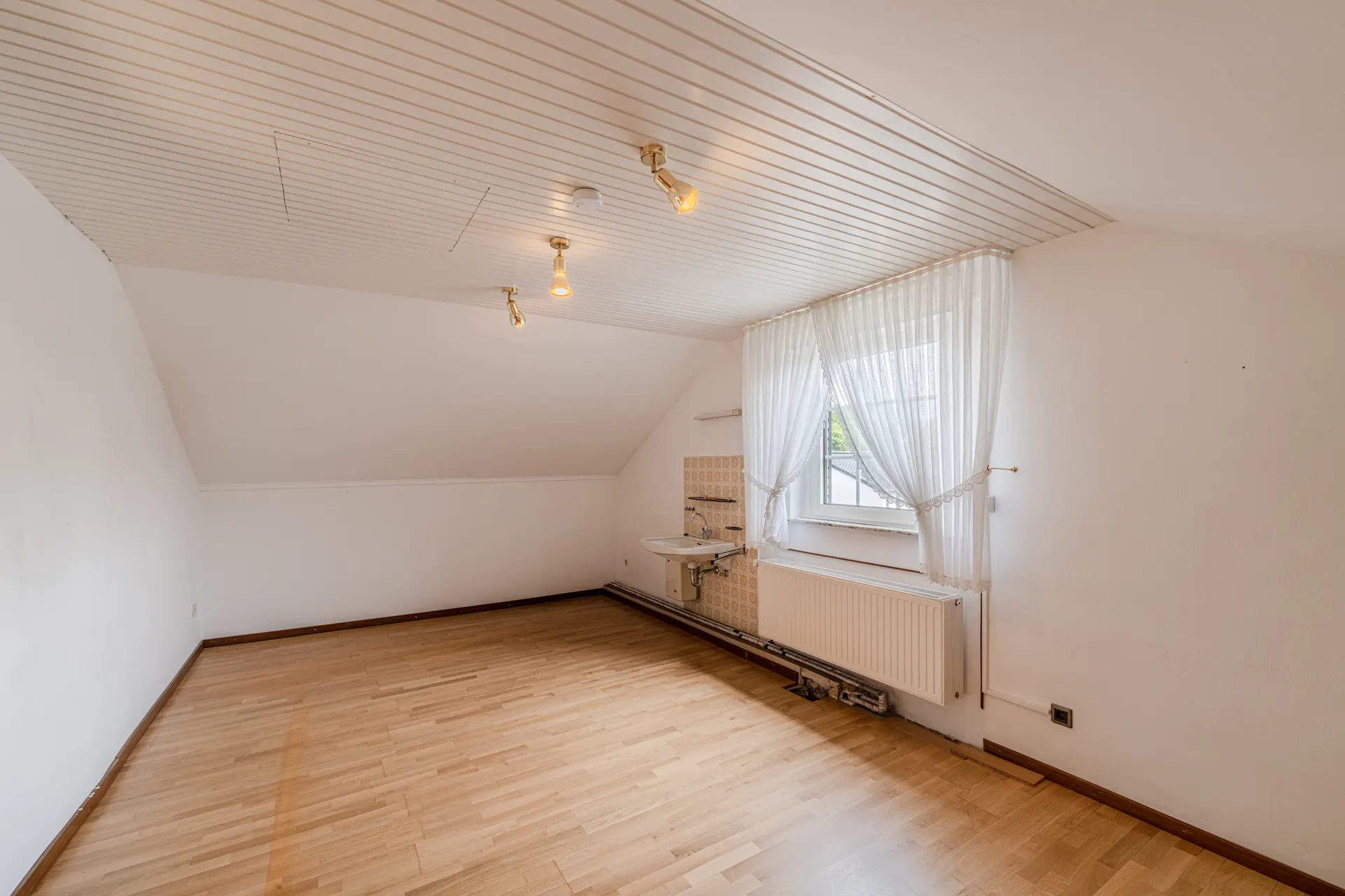 Leeres Zimmer mit Holzboden, Fenster mit Gardinen und Waschbecken an der Wand.