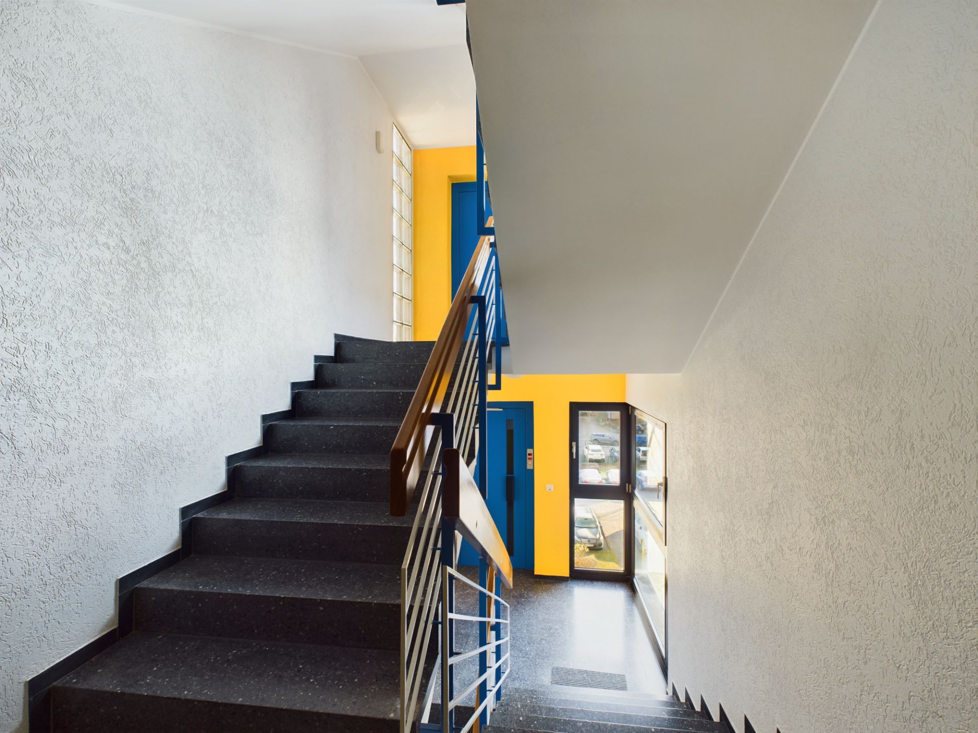 Treppenhaus mit schwarzer Treppe, gelber Wand und blauem Aufzug in einem Wohnhaus.