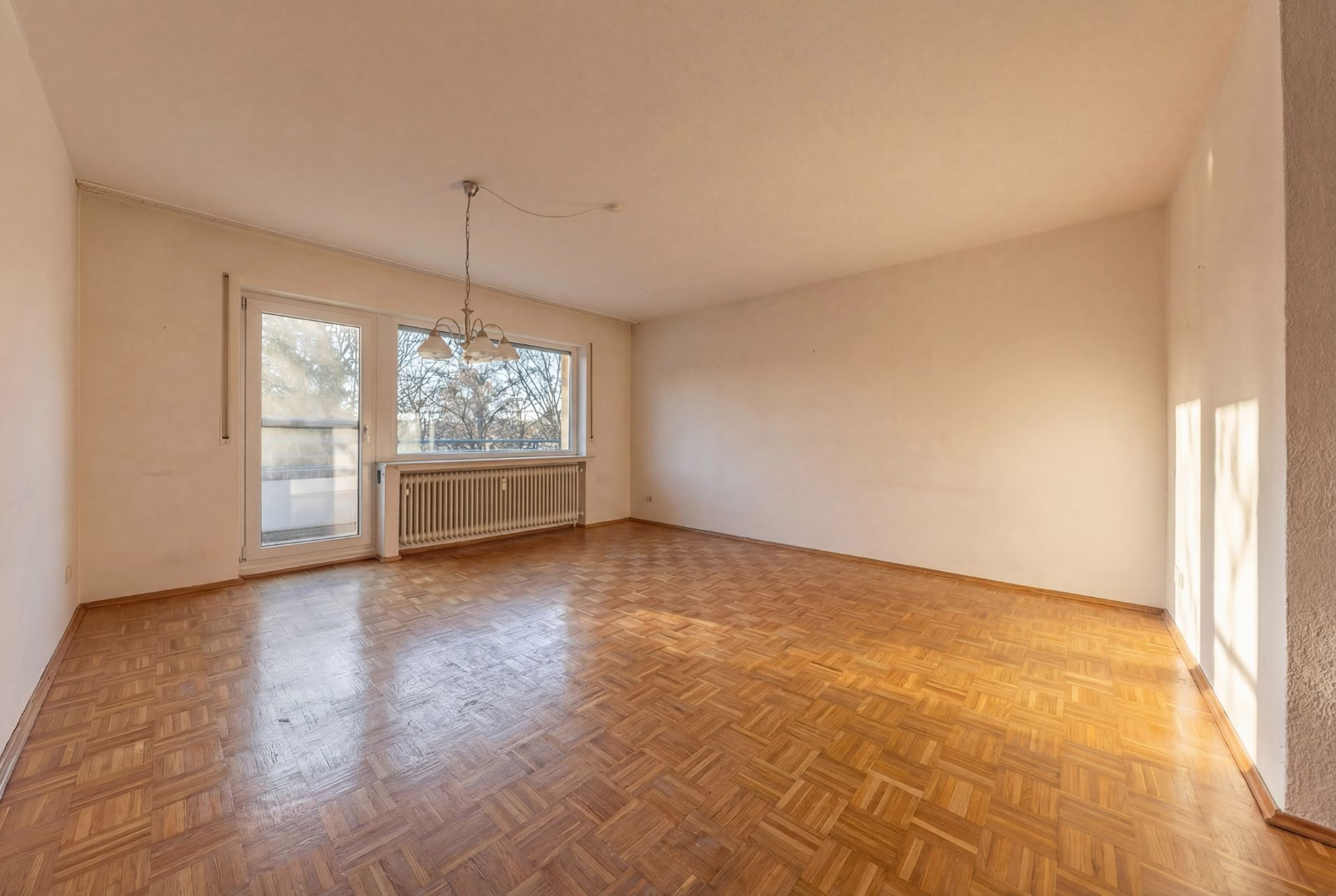 Leerer Raum mit Parkettboden, großen Fenstern und einer Deckenlampe.