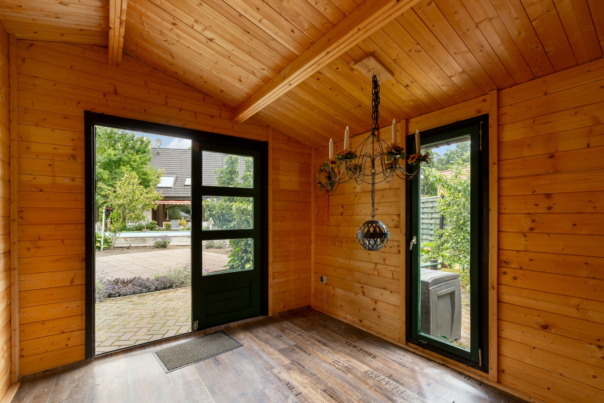 Holzraum mit offenem Fenster, Tür, Kronleuchter und Blick in einen grünen Garten.