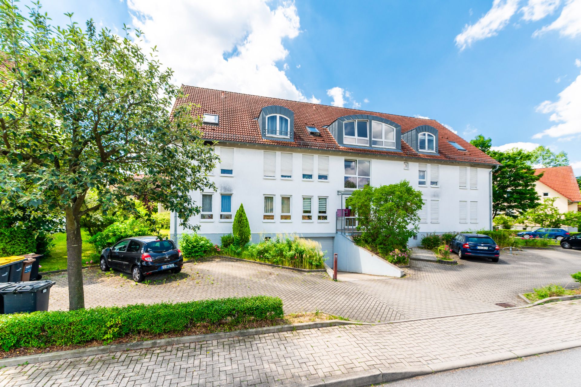 Mehrfamilienhaus mit rotem Dach, umgeben von Bäumen und geparkten Autos auf einem Parkplatz.