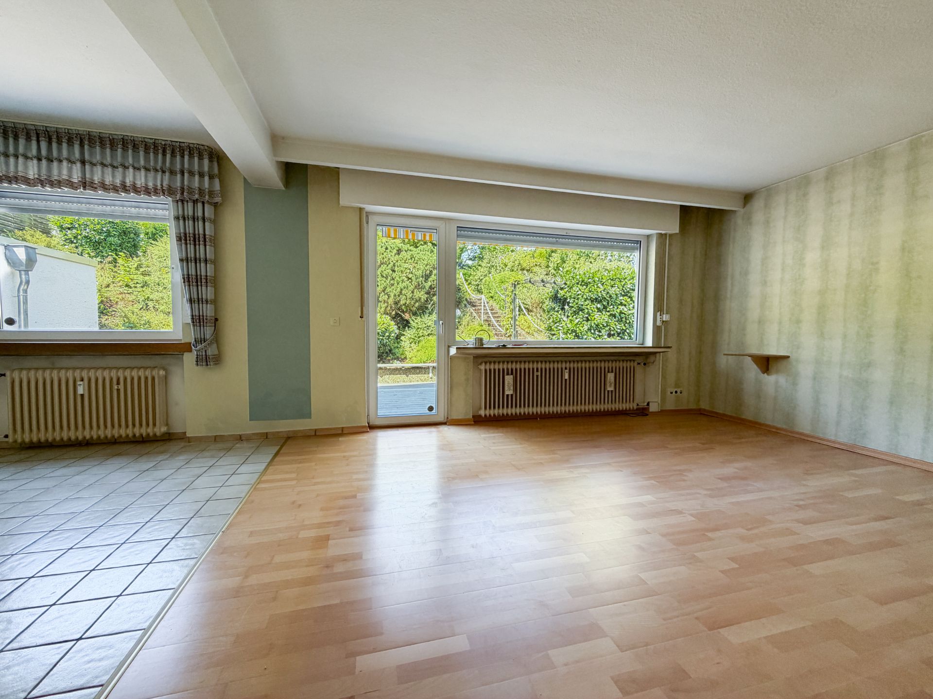 Helles Wohnzimmer mit Parkettboden, großen Fenstern, Heizung und Blick auf einen Garten.