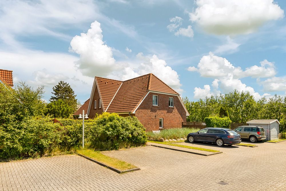 Backsteinwohnhaus mit rotem Dach, Parkplatz und zwei Autos, umgeben von Büschen und Bäumen.