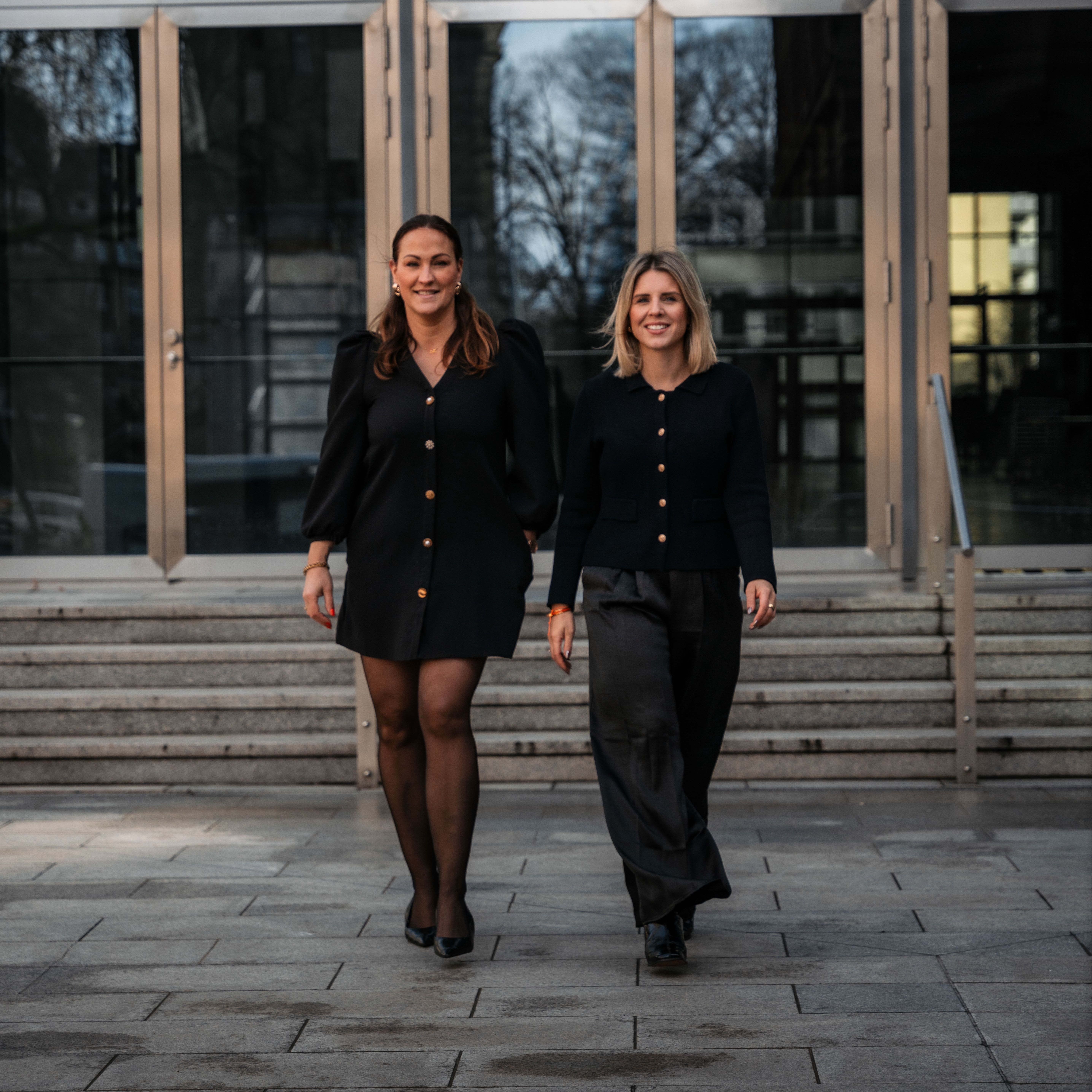 Zwei Frauen in Blazern stehen nebeneinander und lächeln in die Kamera.
