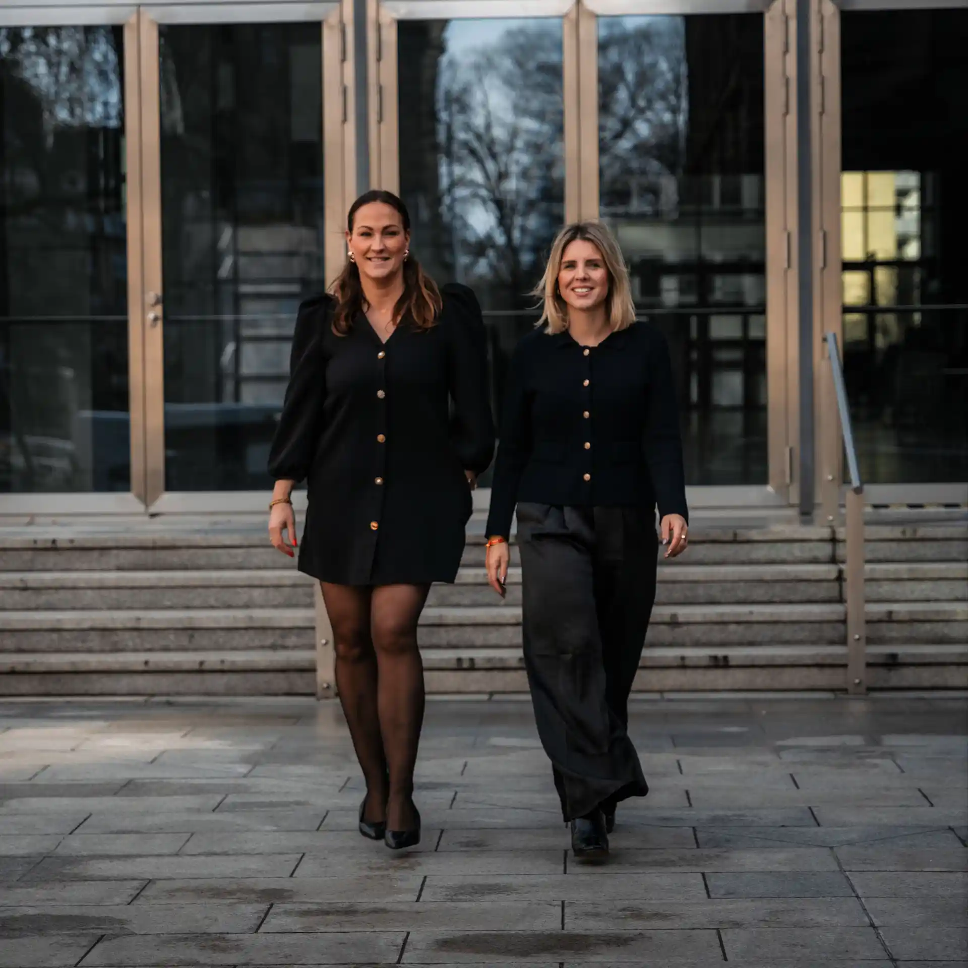 Zwei Frauen in Blazern stehen nebeneinander und lächeln in die Kamera.