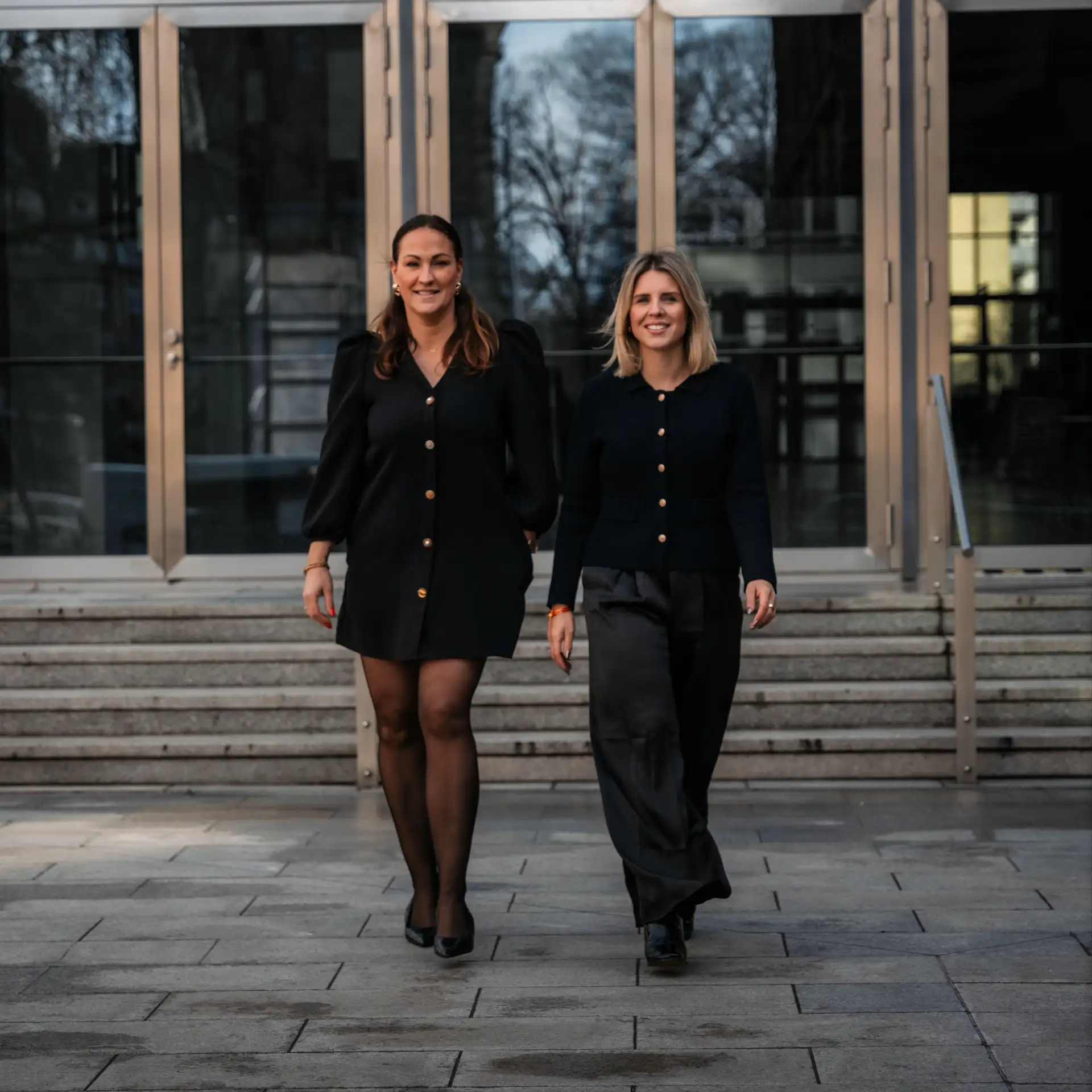 Zwei Frauen in Blazern stehen nebeneinander und lächeln in die Kamera.