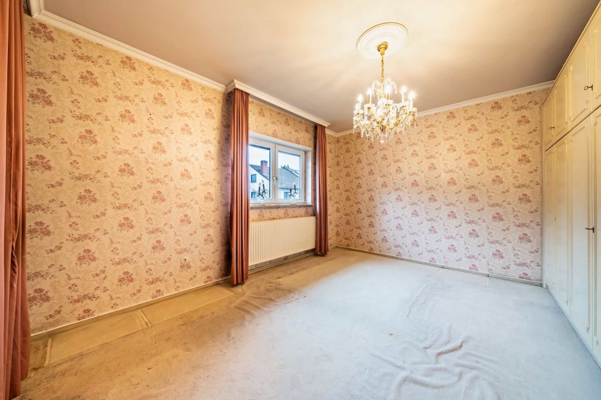 Leeres Zimmer mit Blumenmuster-Tapete, Kronleuchter, Fenster mit Vorhängen und Einbauschrank.
