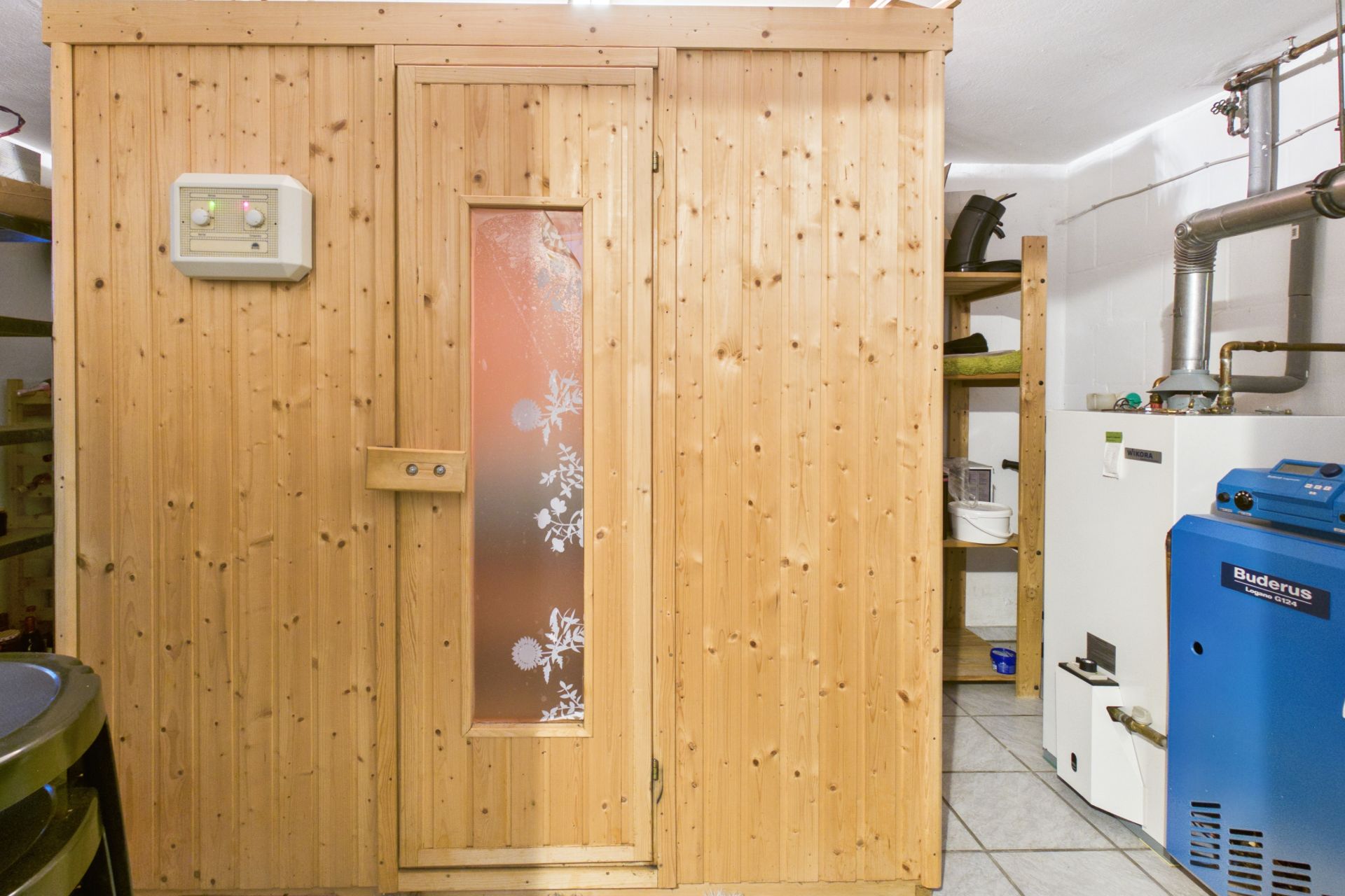 Holzsauna mit Glastür in einem Kellerraum mit Heizungsanlage und Regalen.