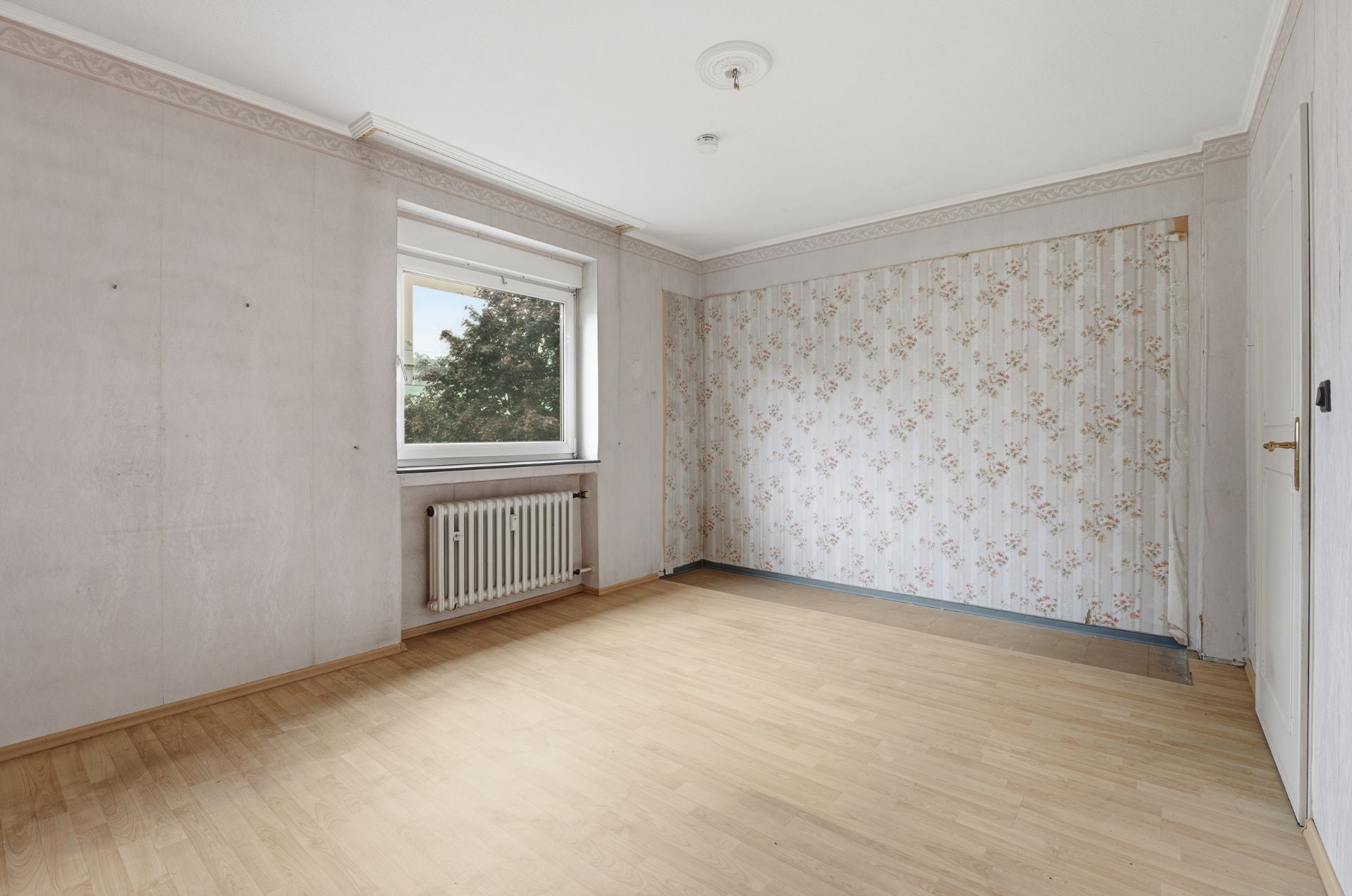 Leeres Zimmer mit Holzboden, Fenster, Heizkörper und gemusterter Tapete an einer Wand.