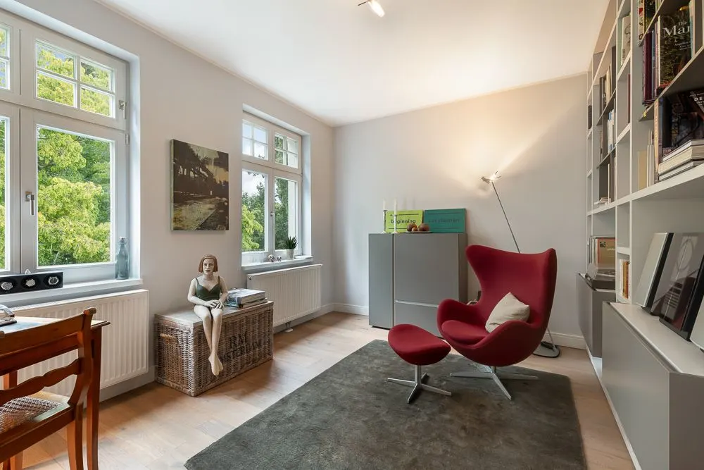 Helles Wohnzimmer mit rotem Sessel, Bücherregal, großen Fenstern und Holzfußboden.