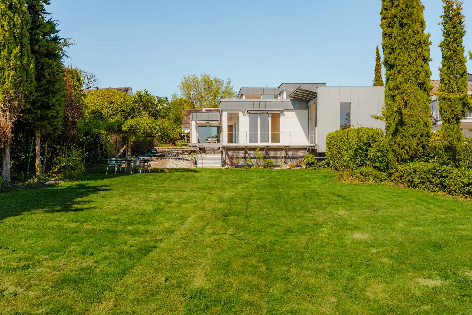 Modernes Haus mit großer grüner Rasenfläche im Garten, umgeben von Bäumen und Büschen.
