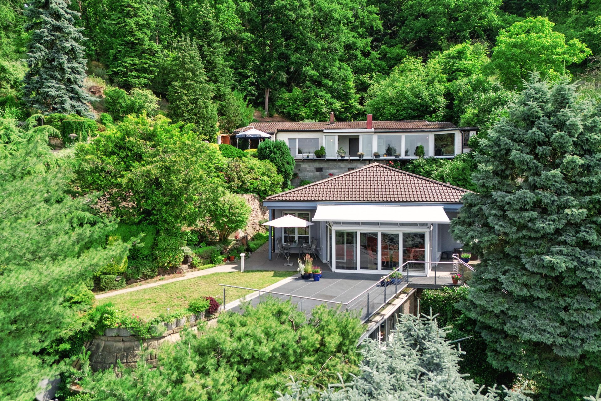 Modernes Haus mit großer Terrasse und Garten, umgeben von vielen grünen Bäumen und Pflanzen.