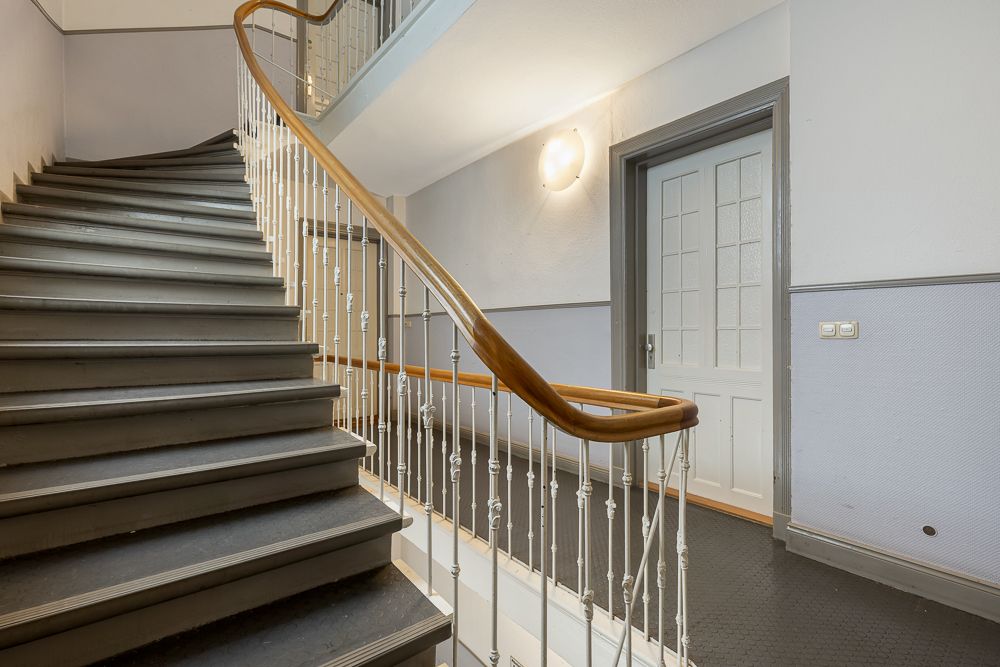 Treppenhaus mit grauer Treppe, Holzgeländer und geschlossener Tür auf dem Absatz.