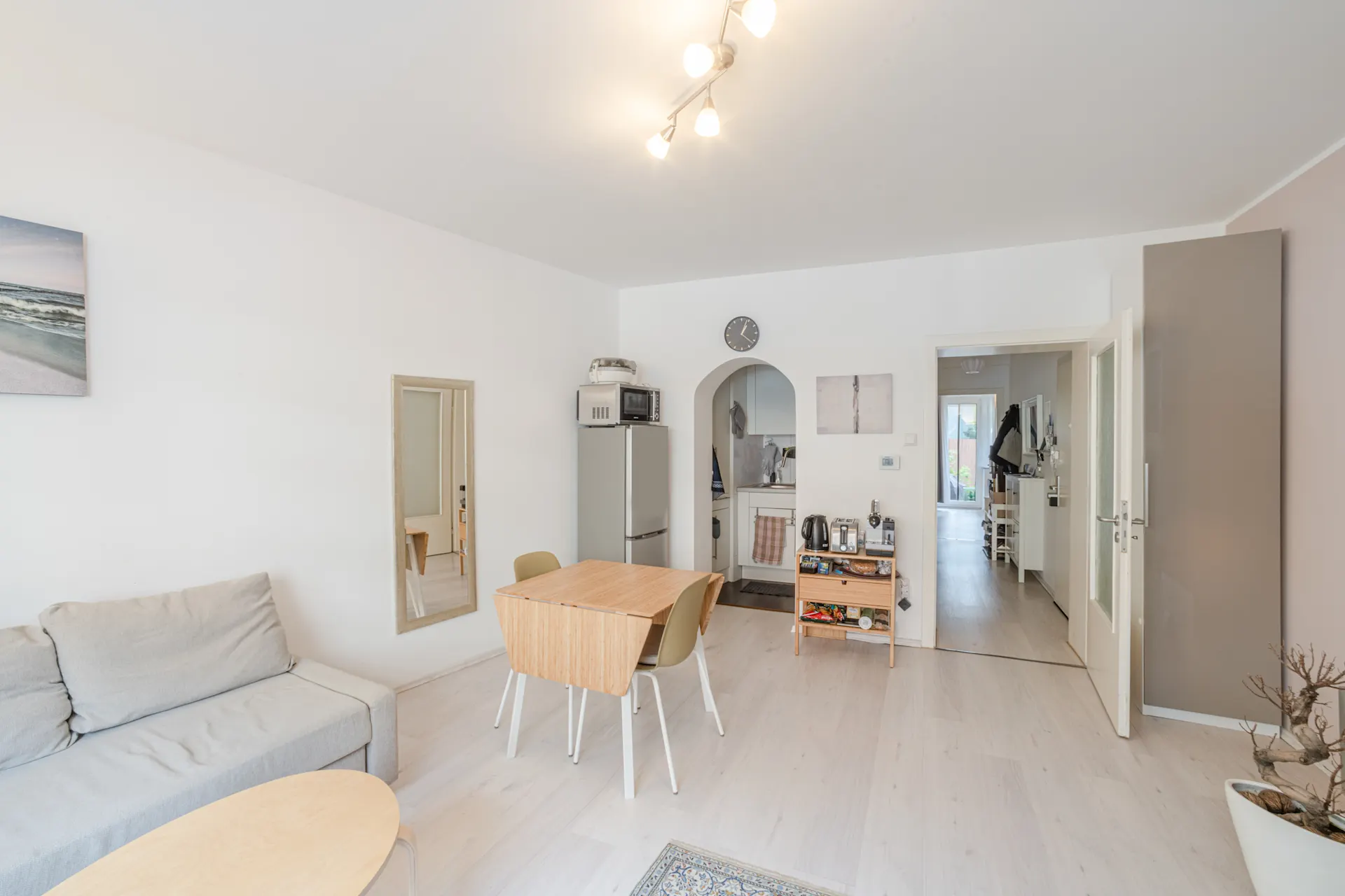Helles Wohnzimmer mit Sofa, Esstisch, Spiegel und offener Küche, Blick in Flur mit Tür zum Garten.