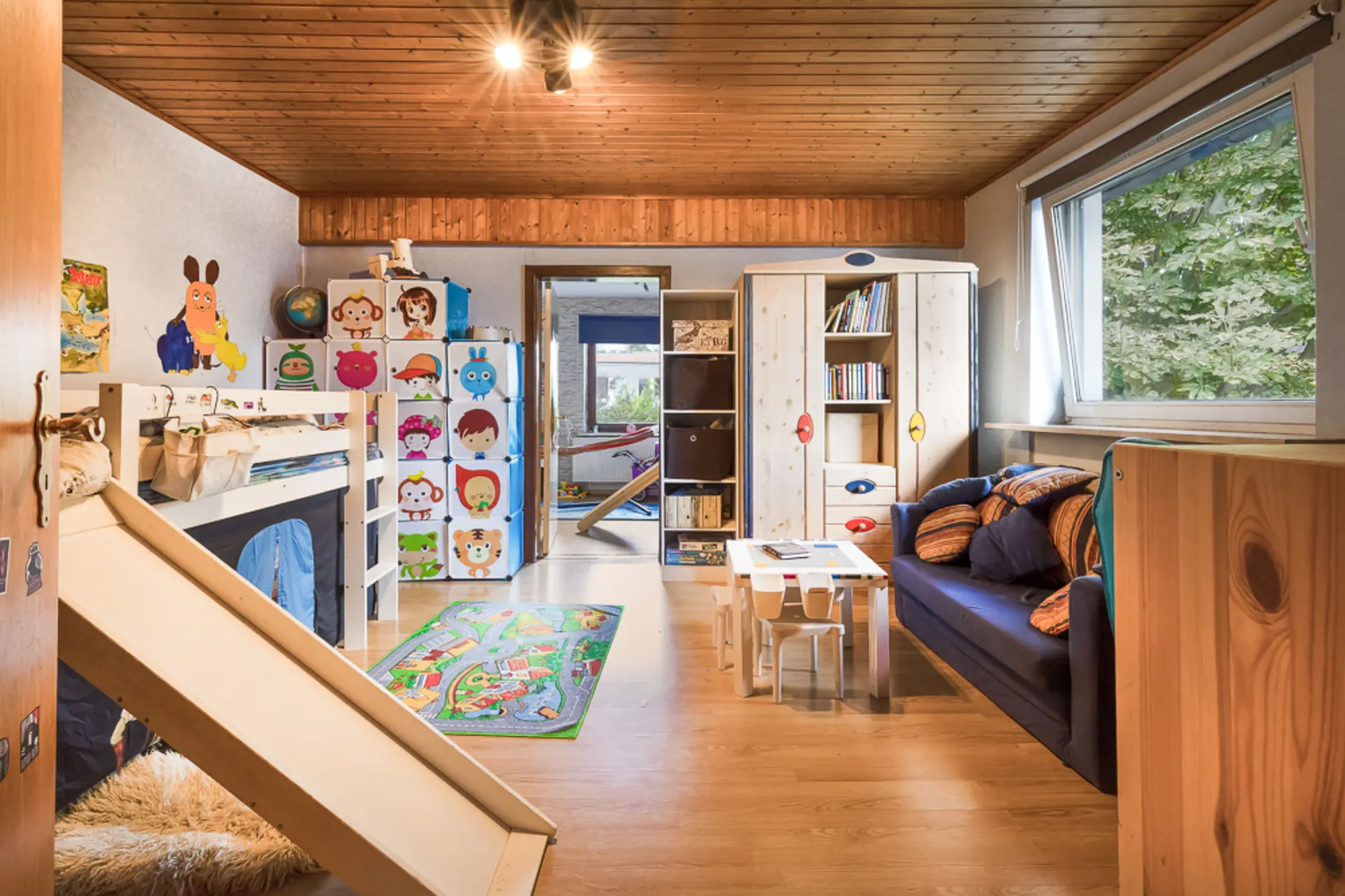 Kinderzimmer mit Etagenbett, Rutsche, bunten Möbeln, Sofa, Tisch und großem Fenster.