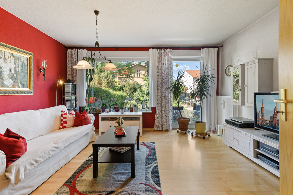 Wohnzimmer mit roter Wand, Sofa, Couchtisch, Fernseher und großen Fenstern mit Blick auf Garten.