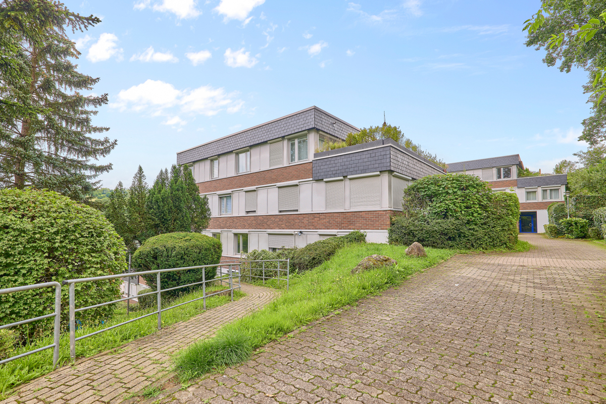 Modernes Wohngebäude mit flachem Dach, großen Fenstern und umgeben von Bäumen und Gehweg.