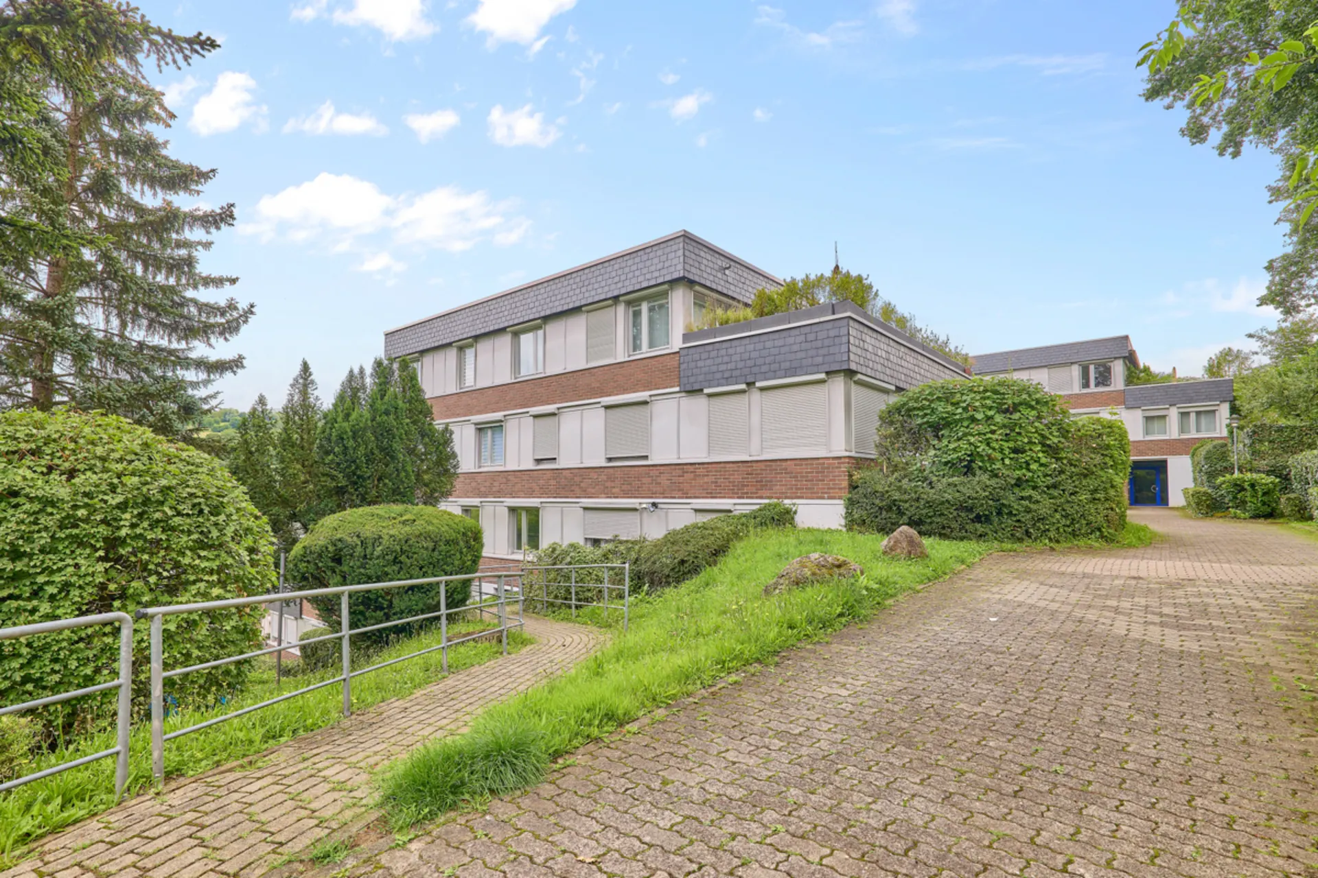 Modernes Wohngebäude mit flachem Dach, großen Fenstern und umgeben von Bäumen und Gehweg.