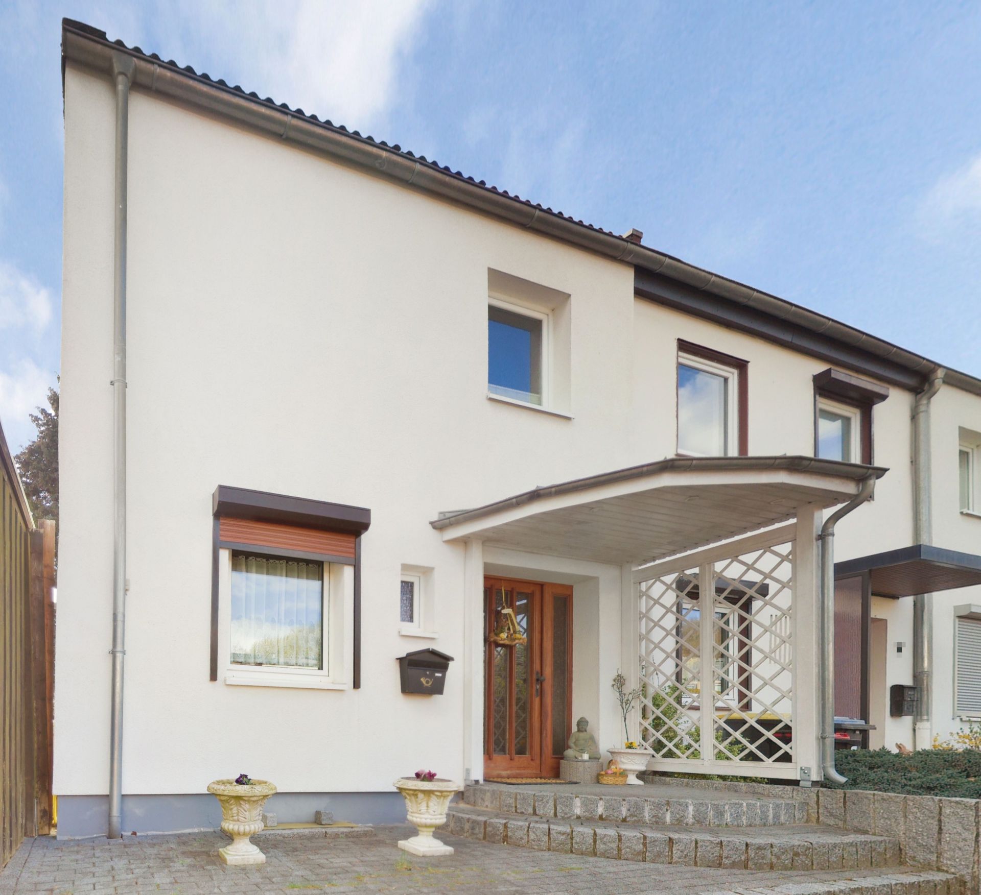 Vorderansicht eines weißen Reihenhauses mit Eingang, überdachter Terrasse und Blumenkübeln.