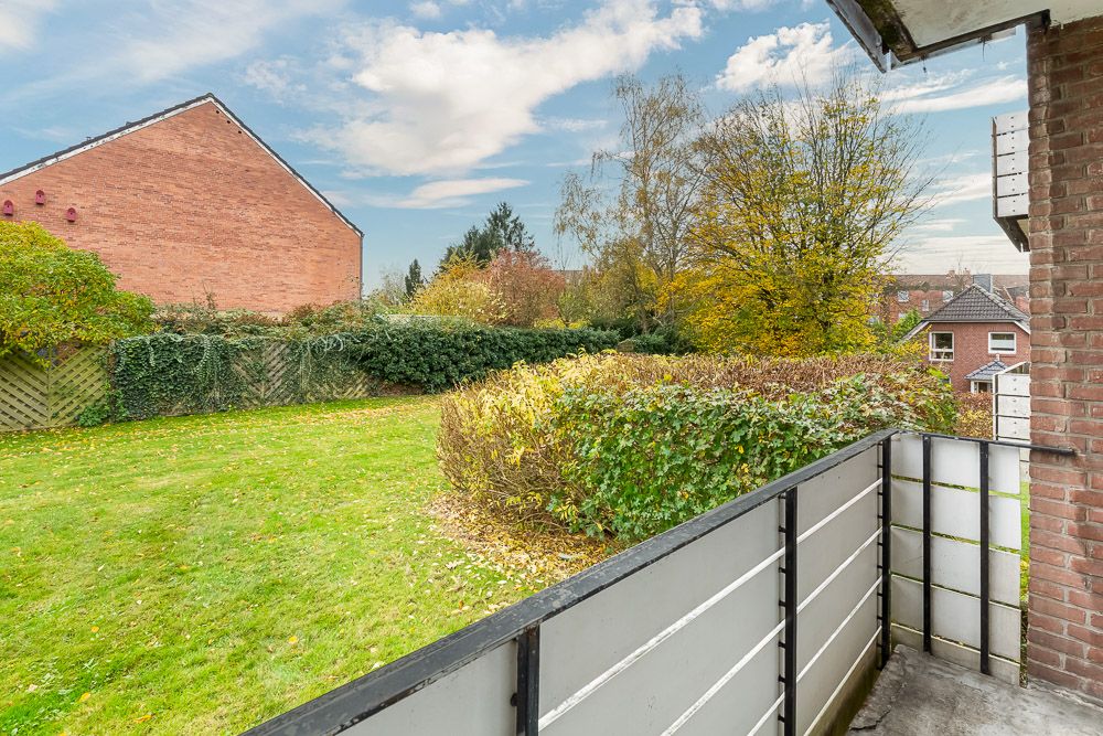Blick von einem Balkon auf einen Garten mit Rasen, Büschen und Bäumen, umgeben von Zäunen und Gebäuden.