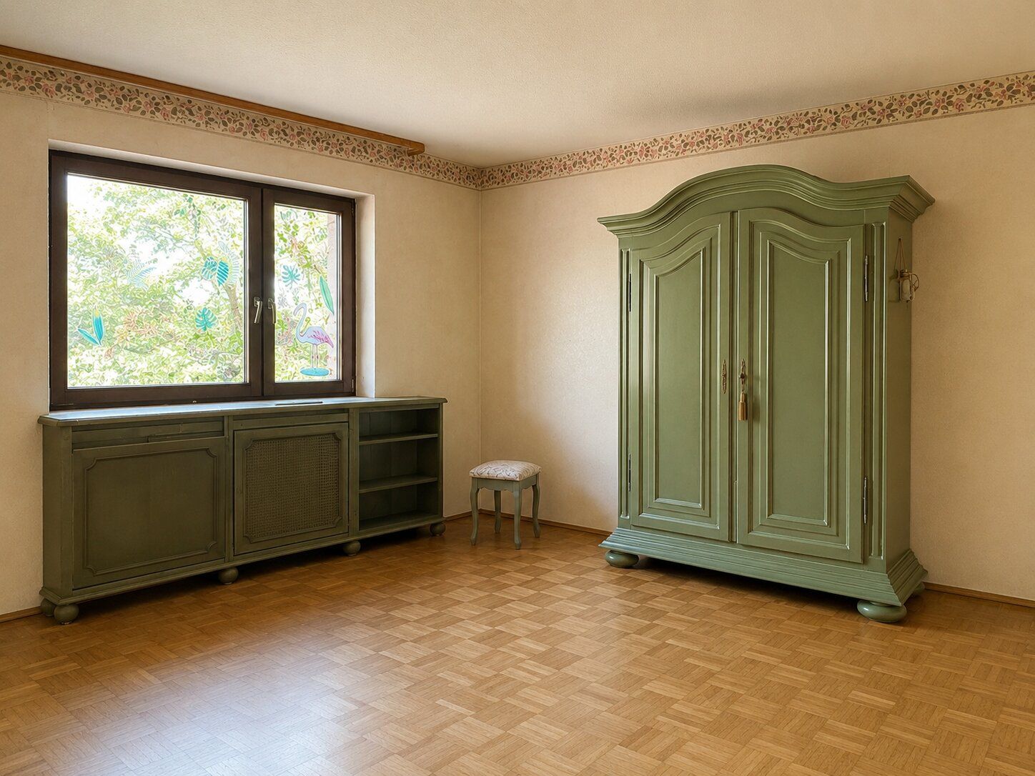 Helles Zimmer mit grünem Schrank, Kommode, kleinem Hocker und großem Fenster.