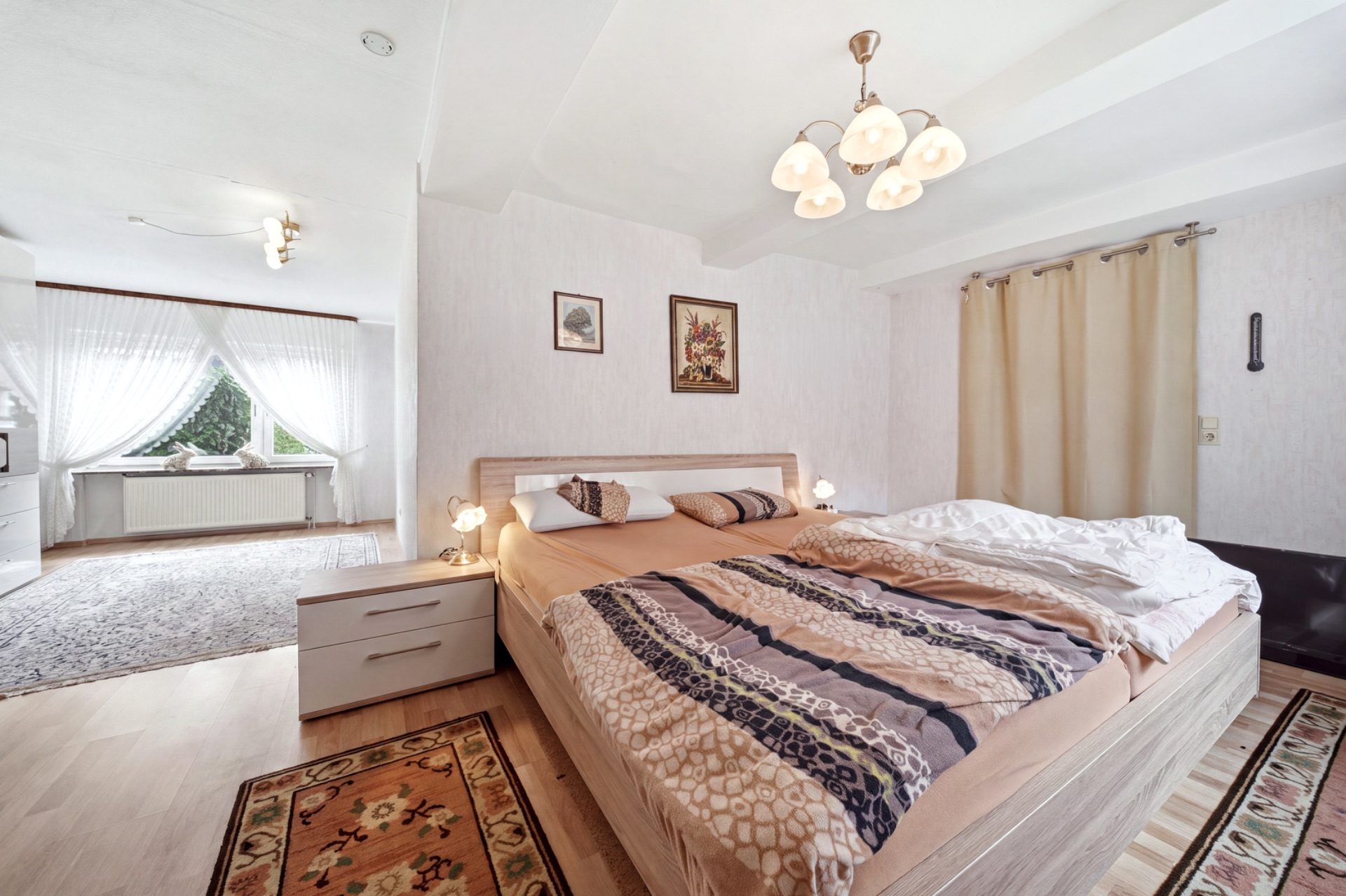 Helles Schlafzimmer mit Doppelbett, Teppichen, Nachttischen und großem Fenster im Hintergrund.