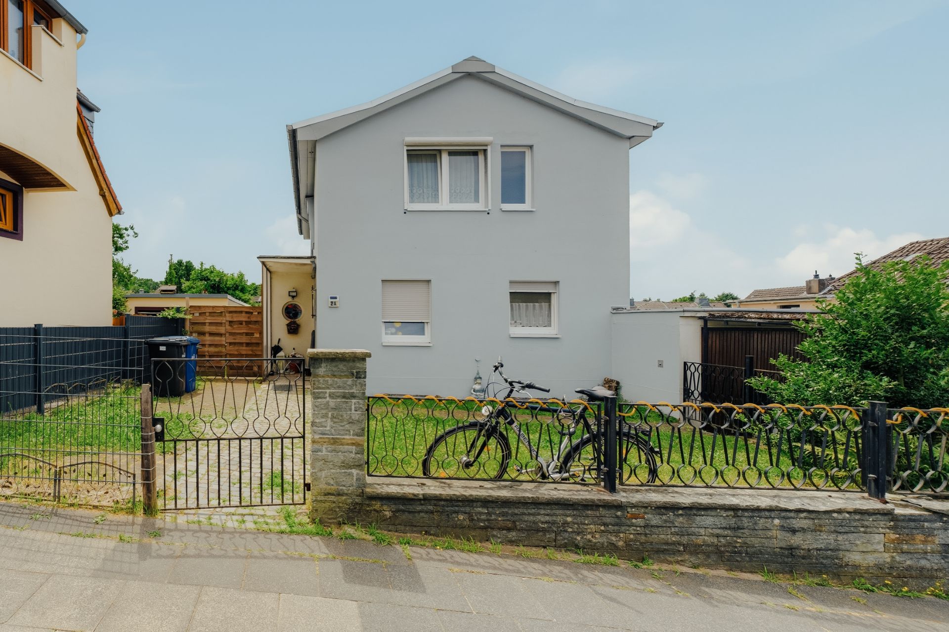 Zweistöckiges Haus mit weißer Fassade, Garten, zwei Fahrrädern und Metallzaun im Vordergrund.