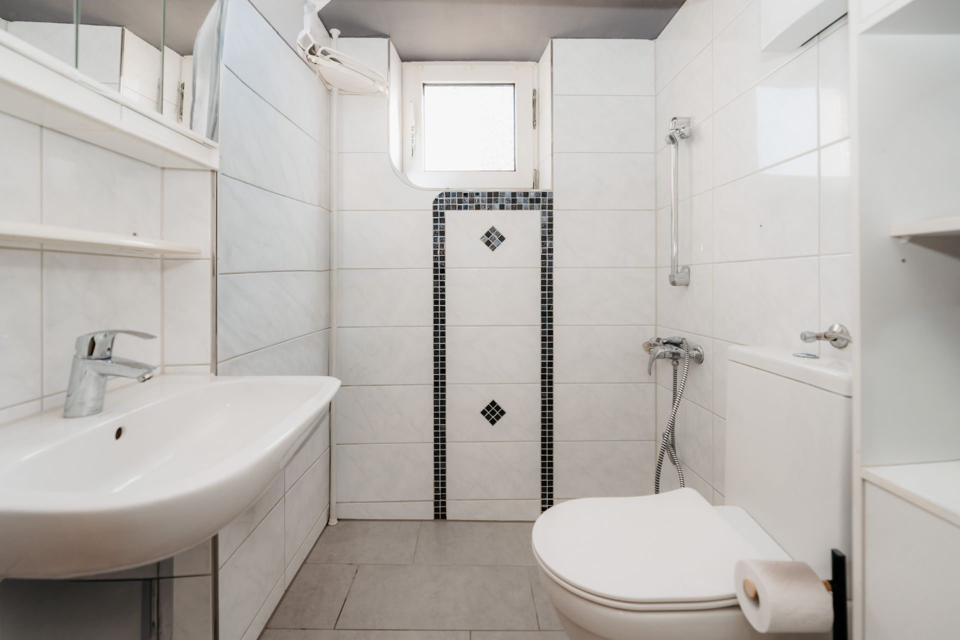 Modernes, weiß gefliestes Badezimmer mit Waschbecken, Toilette und Dusche mit Fenster.