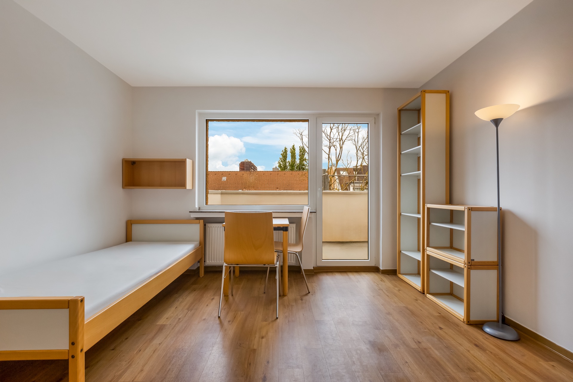 Helles Zimmer mit Einzelbett, Schreibtisch, Stuhl, Regal und Stehlampe, Blick auf Balkon.