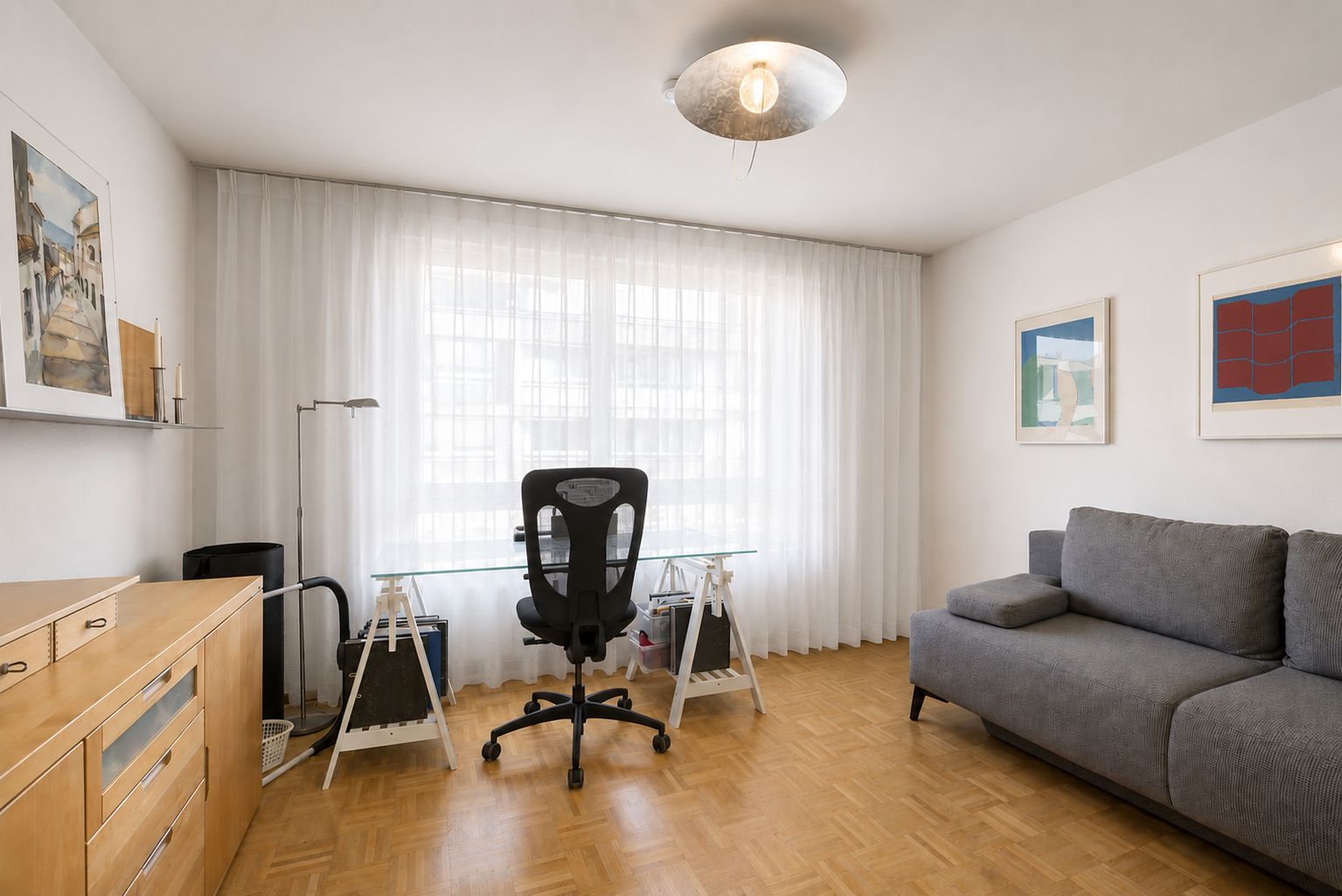 Helles Arbeitszimmer mit Glastisch, Schreibtischstuhl, grauem Sofa und Parkettboden.