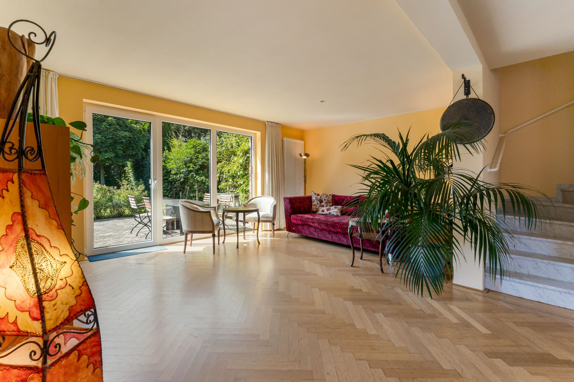 Helles Wohnzimmer mit großer Fensterfront, rotem Sofa, Esstisch, Pflanzen und Zugang zur Terrasse.