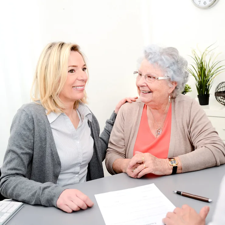 Zwei Frauen sitzen an einem Tisch und sprechen mit einer dritten Person, Dokumente liegen auf dem Tisch.