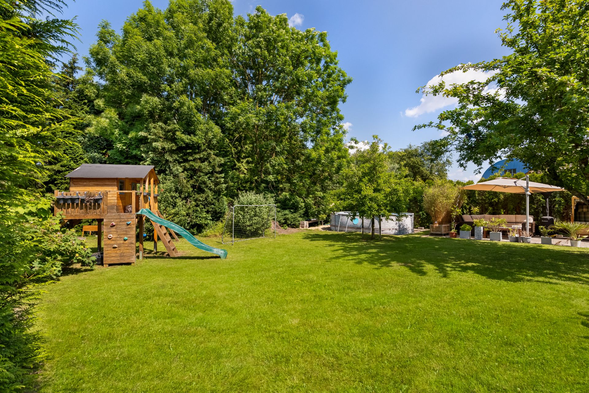 Großer Garten mit Spielhaus, Rutsche, Sitzbereich unter Sonnenschirm und viel Grün.