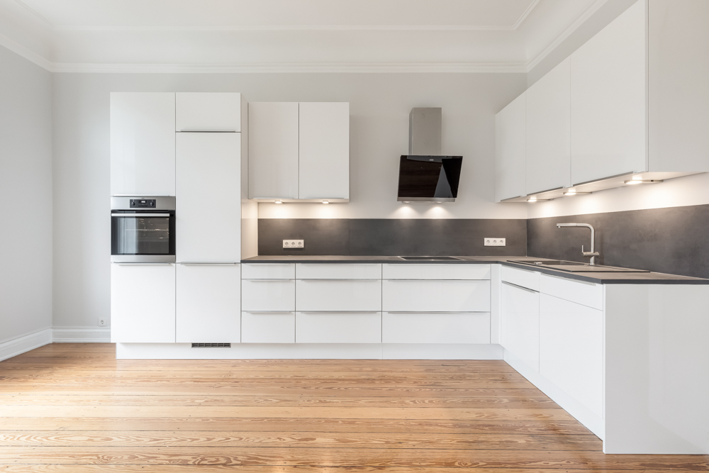 Moderne, weiße Einbauküche mit Holzfußboden, Backofen und grauer Arbeitsplatte.
