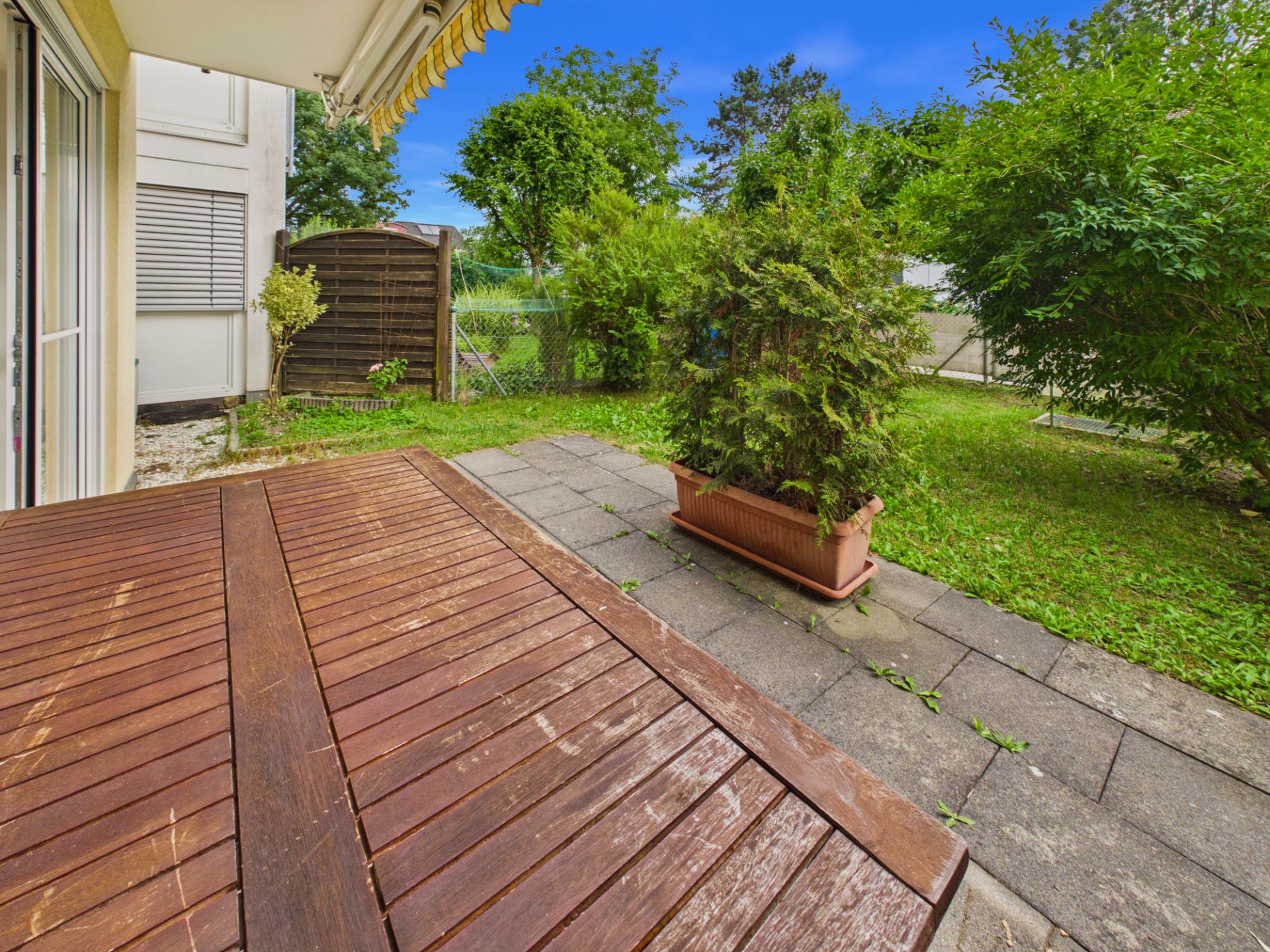 Terrasse mit Holztisch, Pflanzkübel und Garten mit Rasen und Bäumen.