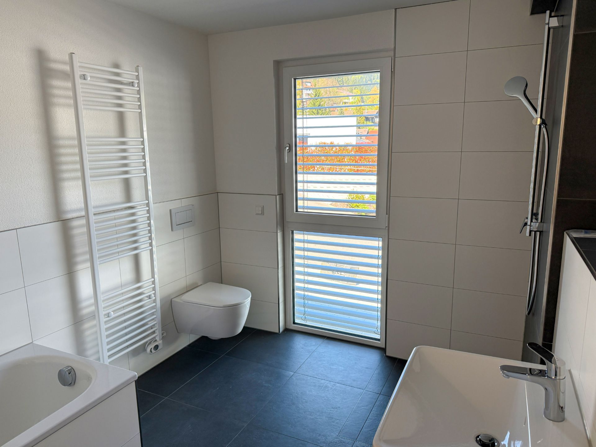 Modernes Badezimmer mit Badewanne, Dusche, WC, Handtuchheizkörper und Fenster mit Jalousie.