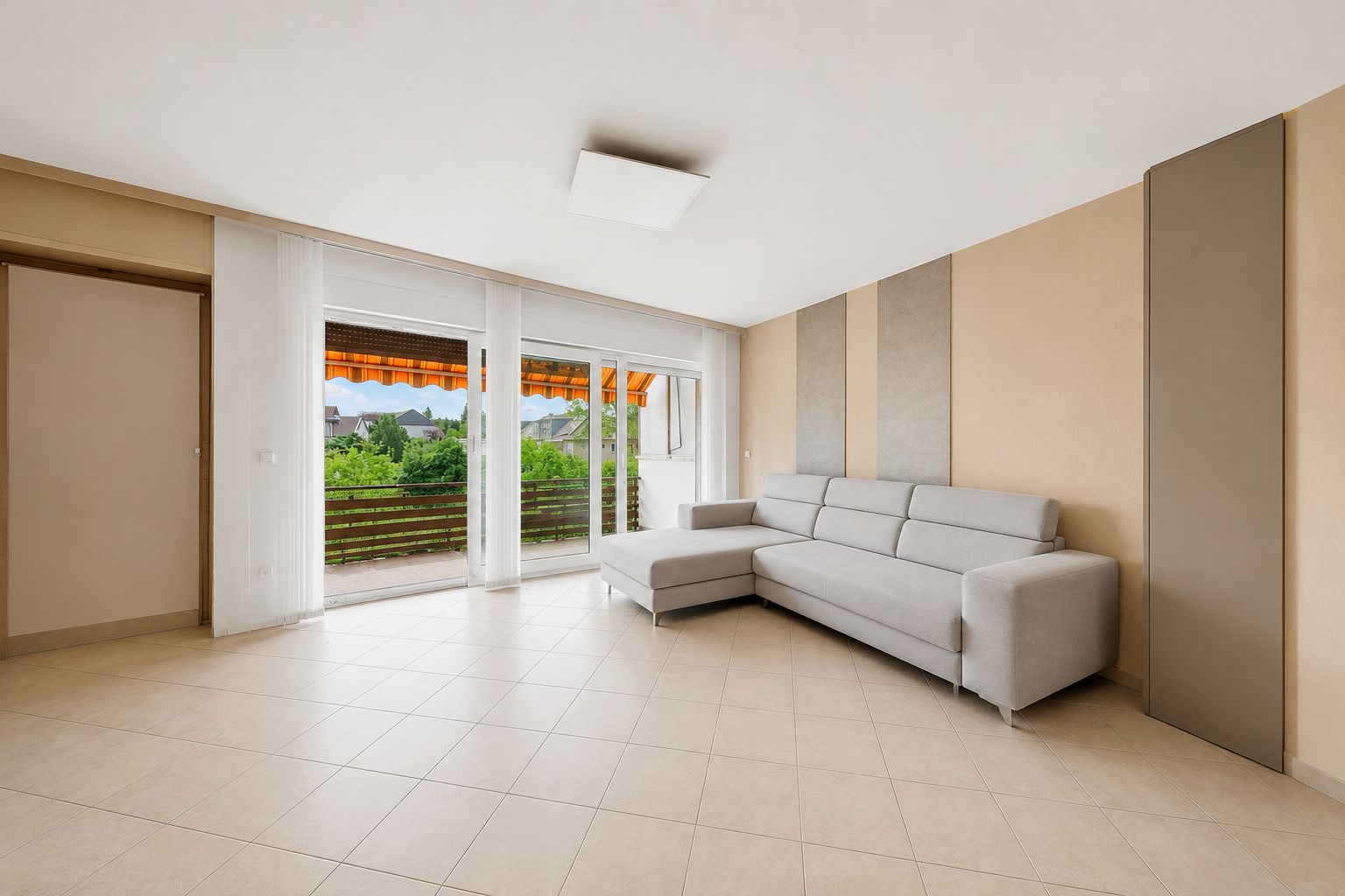 Helles Wohnzimmer mit grauem Ecksofa, Fliesenboden und großer Fensterfront zum Balkon.
