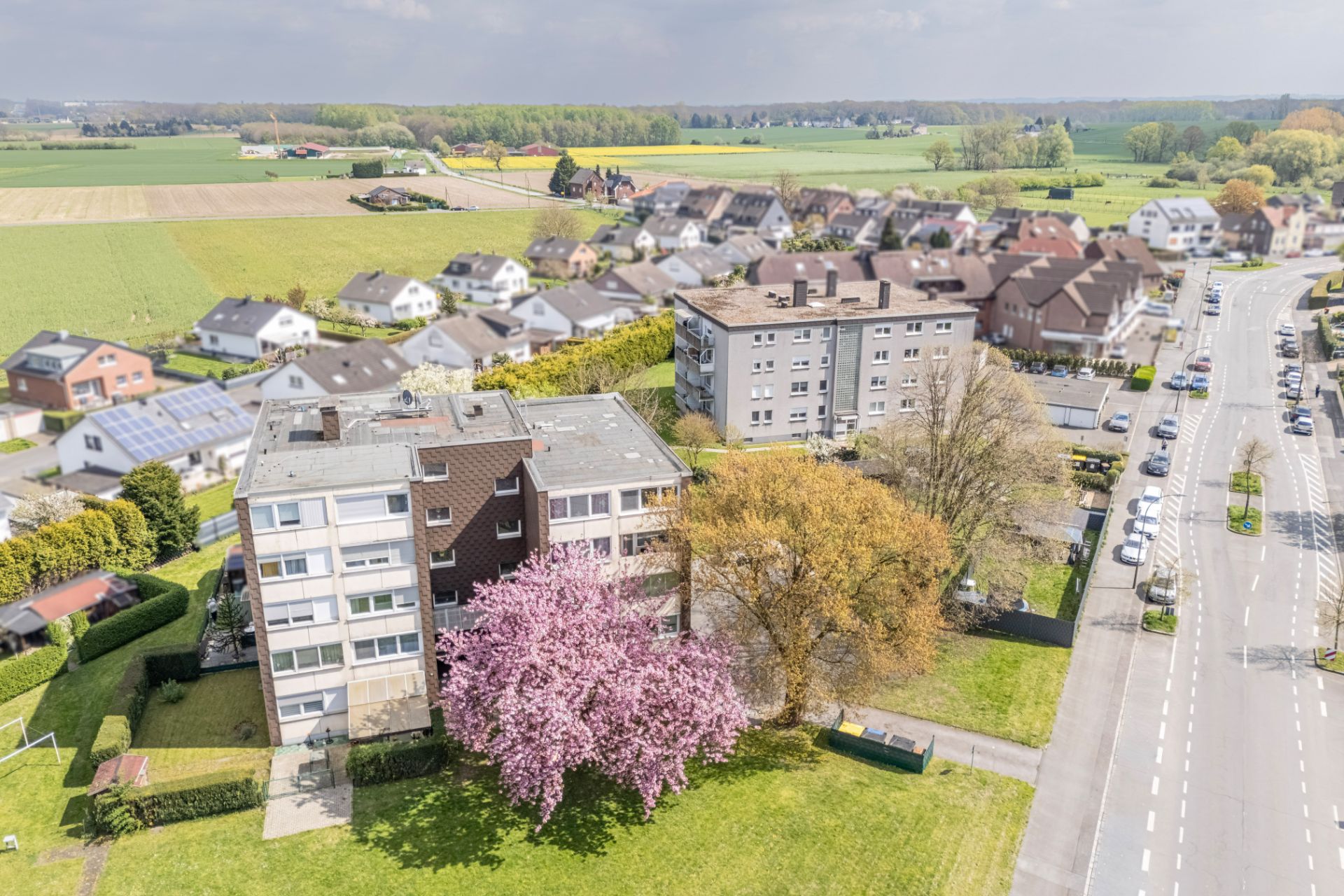 Mehrfamilienhaus mit blühendem Baum davor, daneben weitere Gebäude und eine Straße.