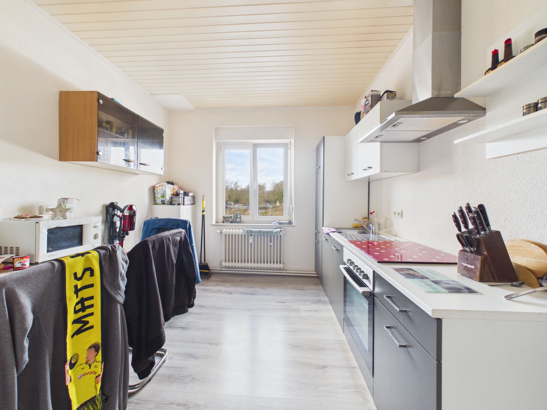 Helle, schmale Küche mit Herd, Kühlschrank, Esstisch und Fenster mit Blick nach draußen.