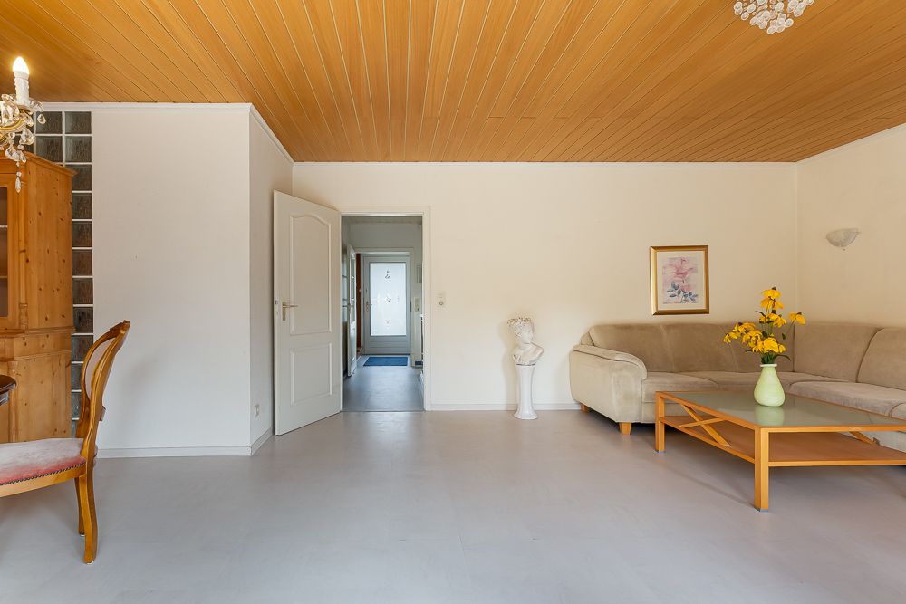 Helles Wohnzimmer mit Ecksofa, Glastisch, Blumenstrauß und Holzdecke.