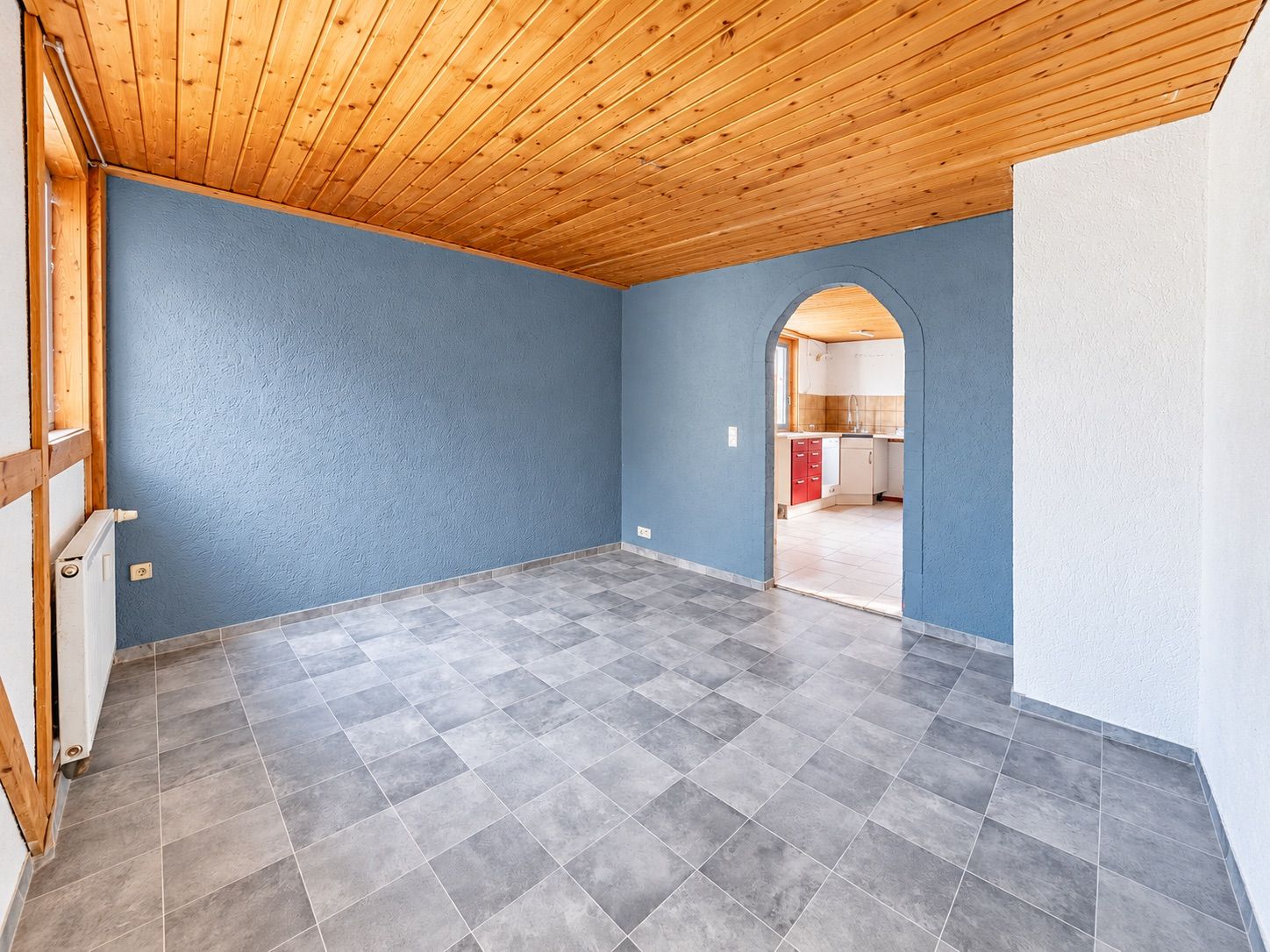 Leeres Zimmer mit blauer Wand, Holzdecke und Durchgang zur Küche.