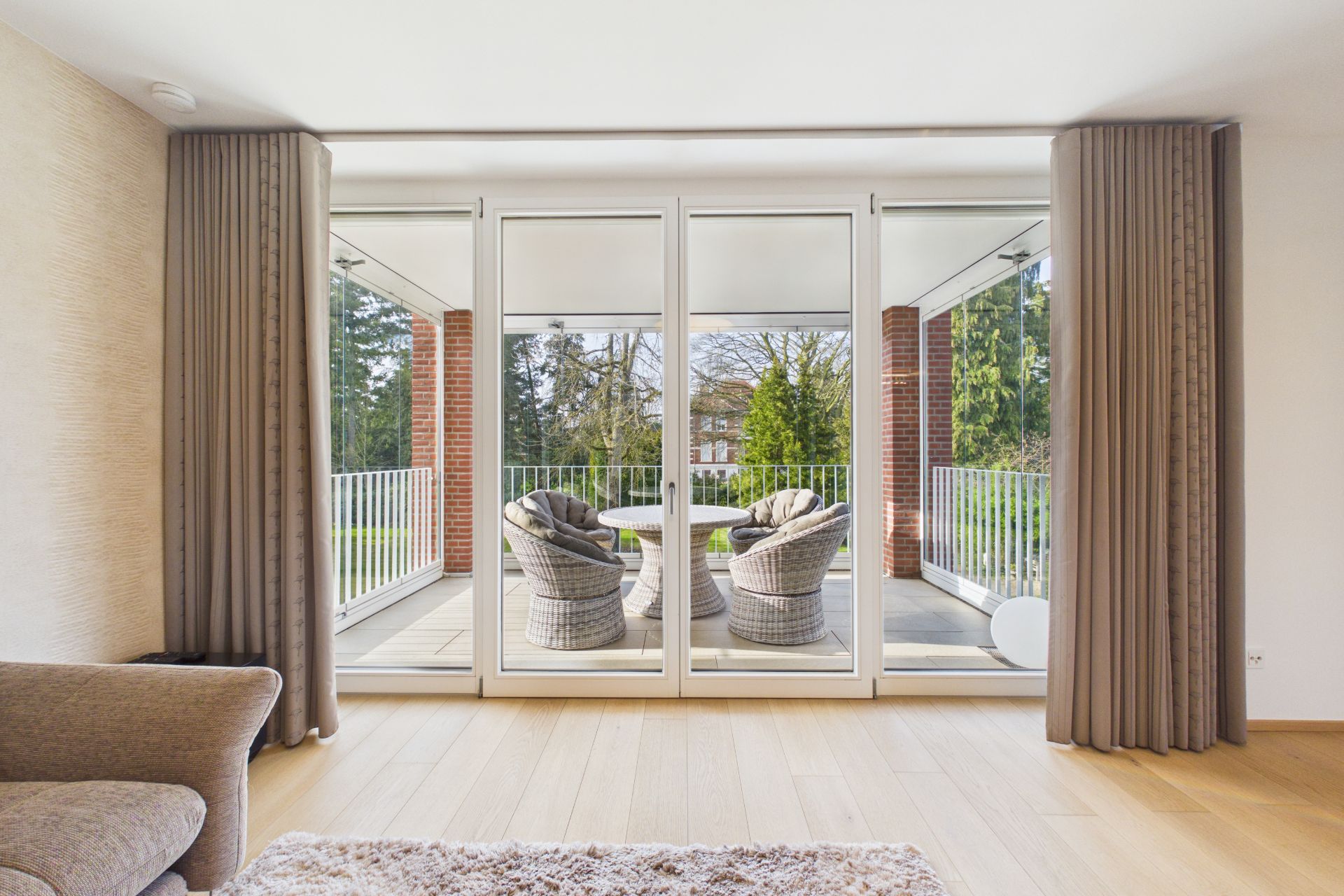 Wohnzimmer mit großen Fenstern und Blick auf einen Balkon mit vier Rattan-Sesseln und Gartenaussicht.
