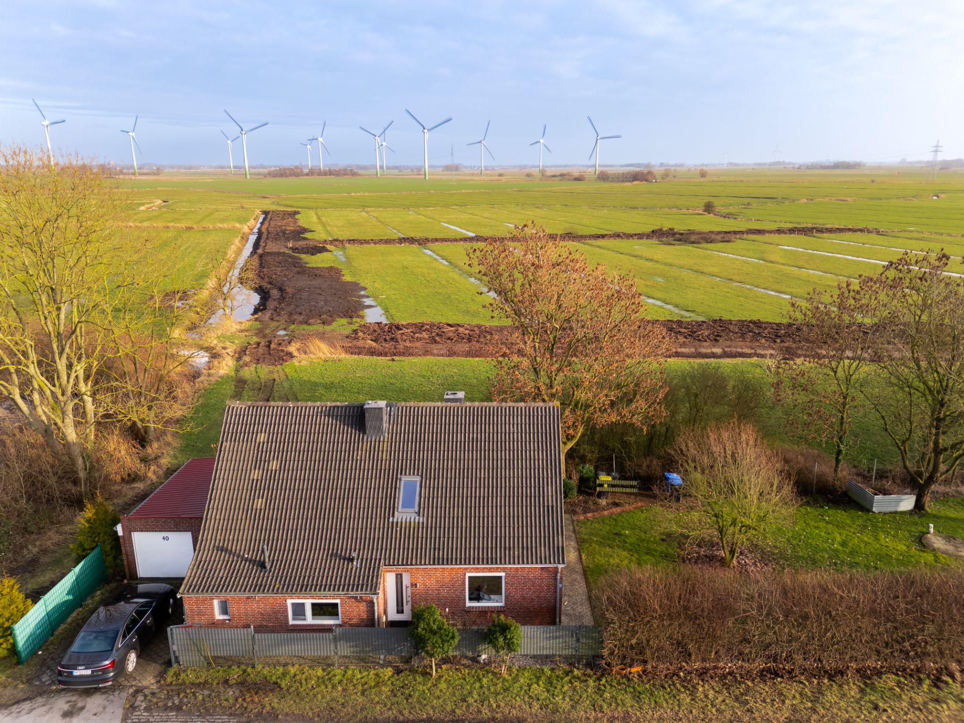 Backsteinwohnhaus mit Garten und Feldern im Hintergrund, Windräder in der Ferne.