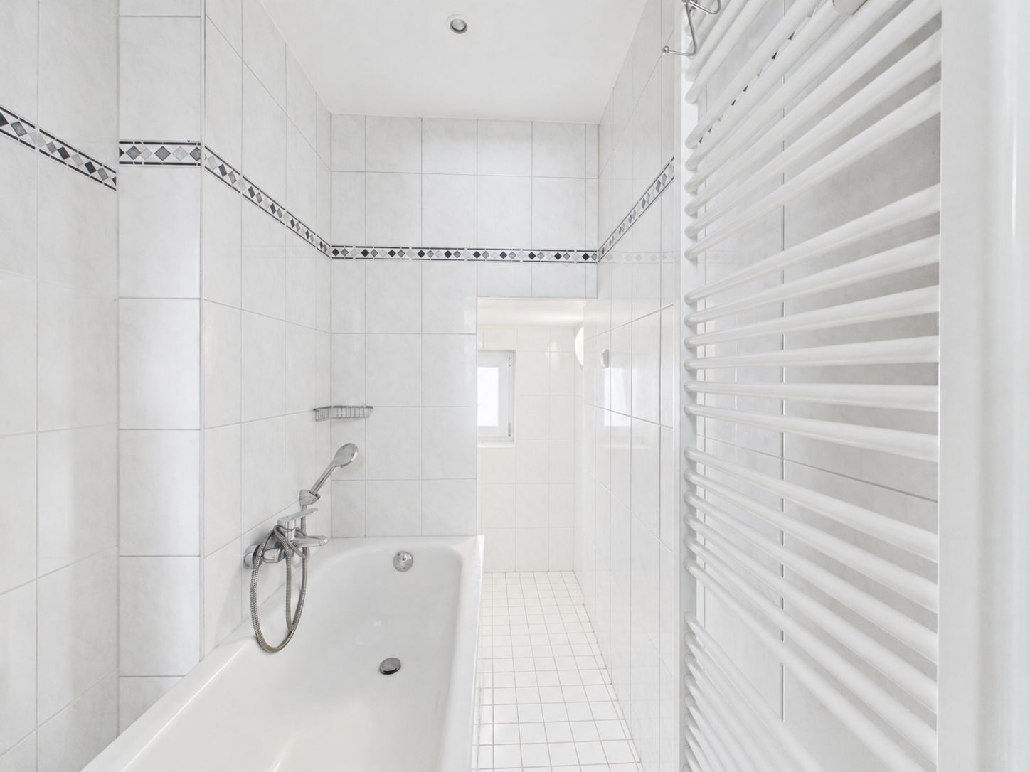 Helles Badezimmer mit Badewanne, weiß gefliest, Heizkörper und kleinem Fenster am Ende.