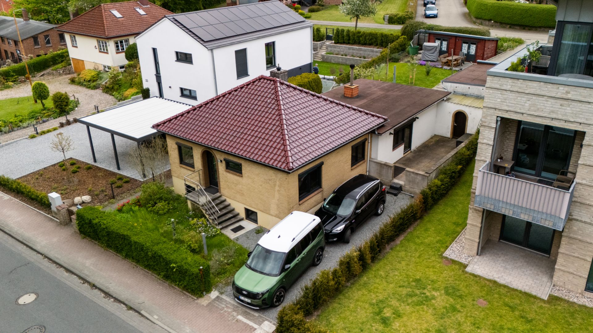 Luftaufnahme eines Einfamilienhauses mit rotem Dach, zwei Autos in der Einfahrt und umliegendem Garten.